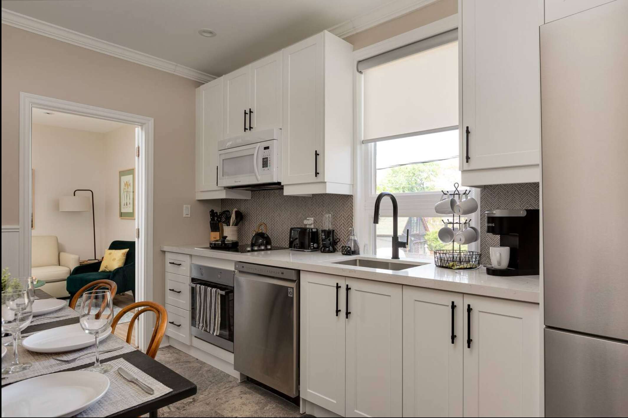 Modern kitchen with white shaker cabinets, light quartz countertops, stainless steel appliances including a dishwasher and microwave, and a herringbone tile backsplash. A dining table with place settings is visible in the foreground.