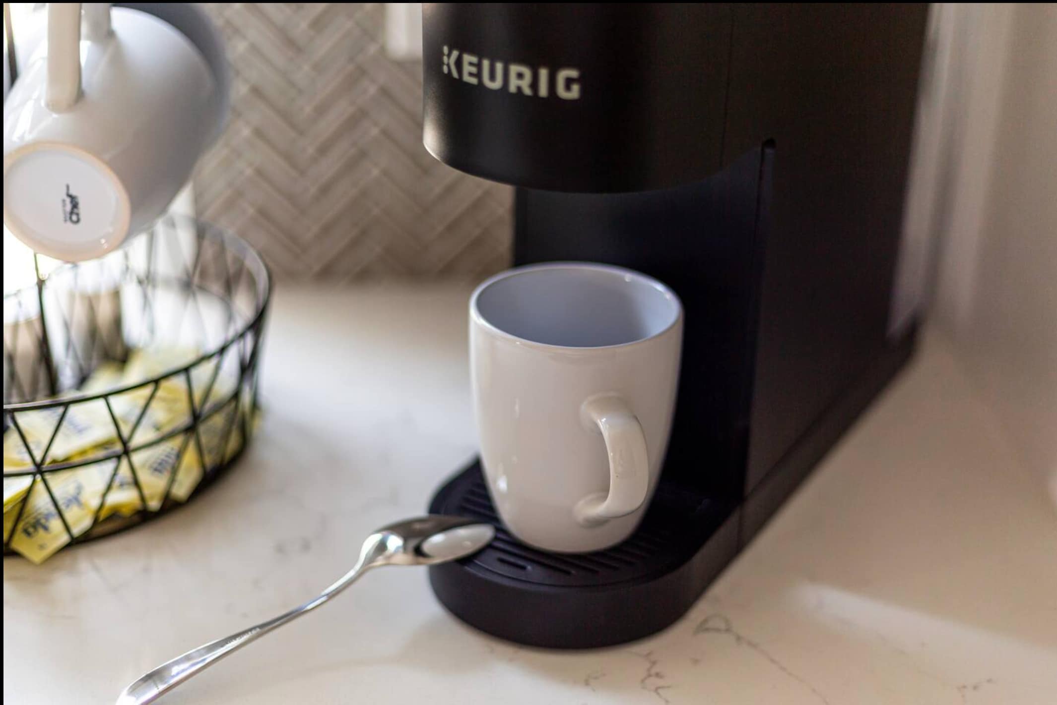 A close-up of a black Keurig coffee maker on a white countertop, with a white mug ready for brewing. A metal basket with coffee pods and a spoon are also visible.