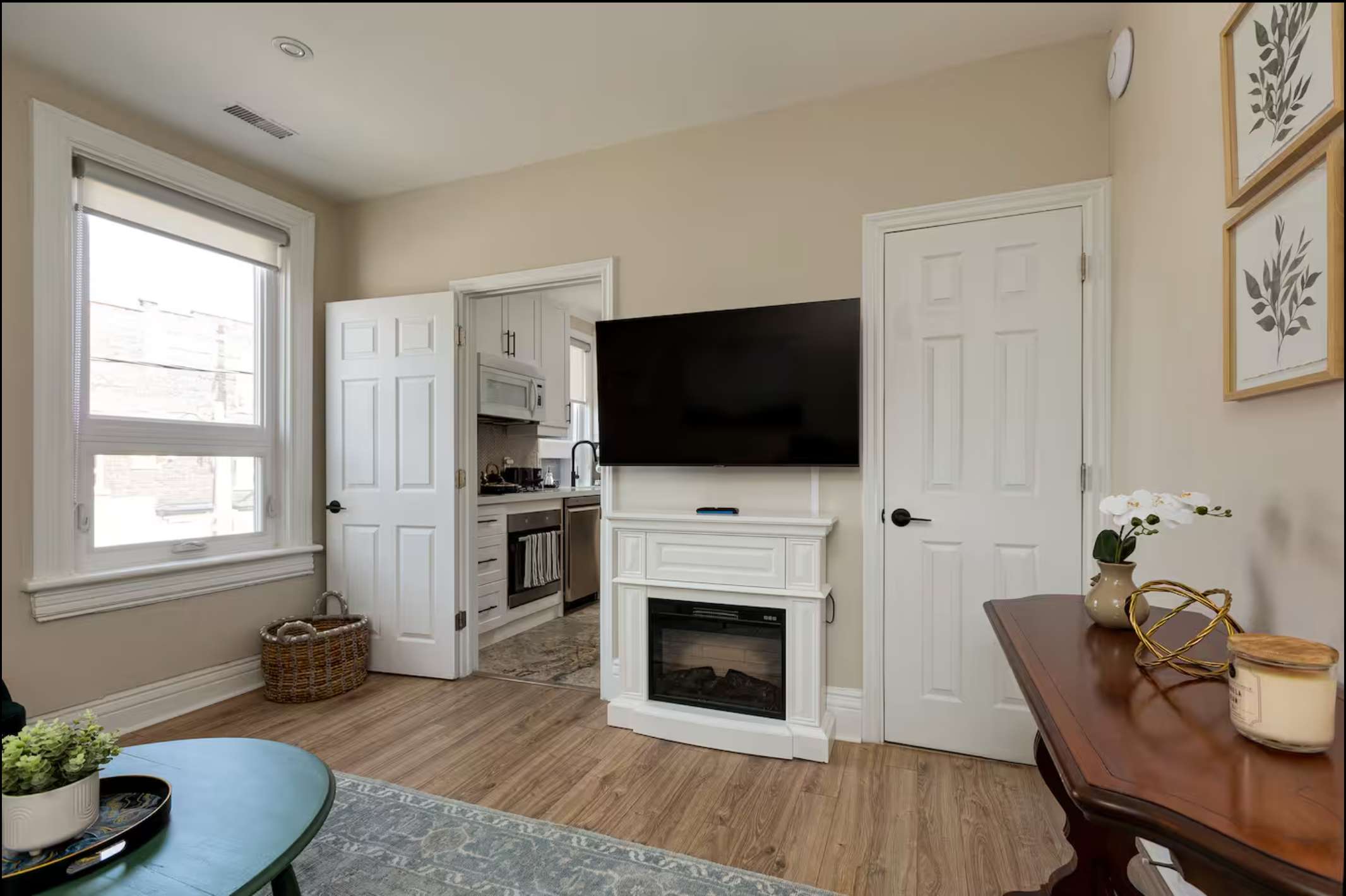 The living room features a wall-mounted TV above a white electric fireplace, with a doorway leading to the kitchen. A round green coffee table and a patterned rug define the seating area.