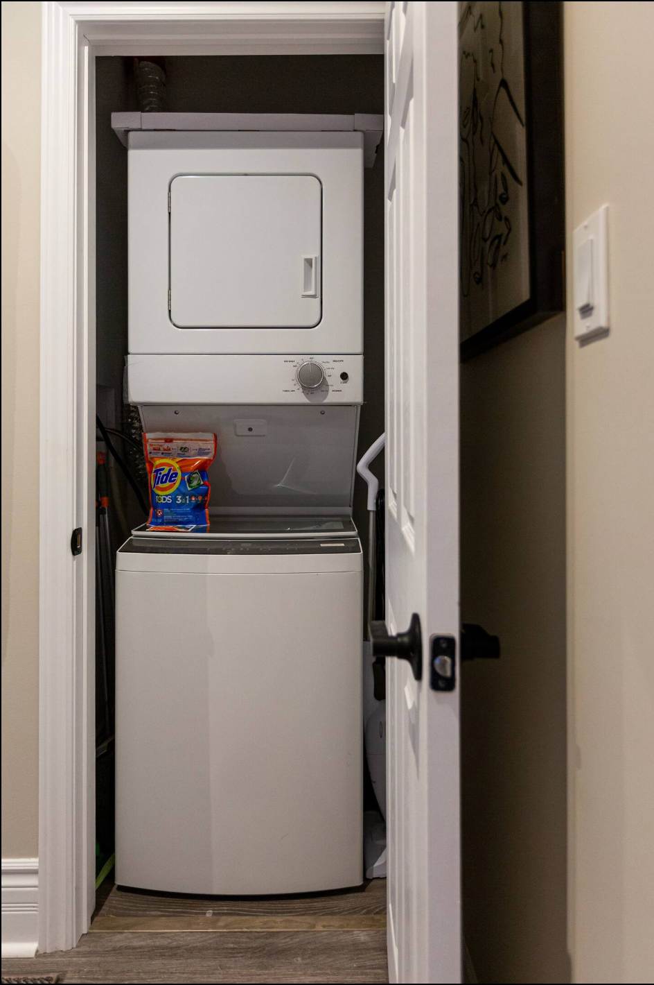 Compact laundry closet featuring a white stacked washer and dryer unit, with a bag of Tide Pods on top of the washer. Cleaning supplies are visible to the left and right of the appliances.