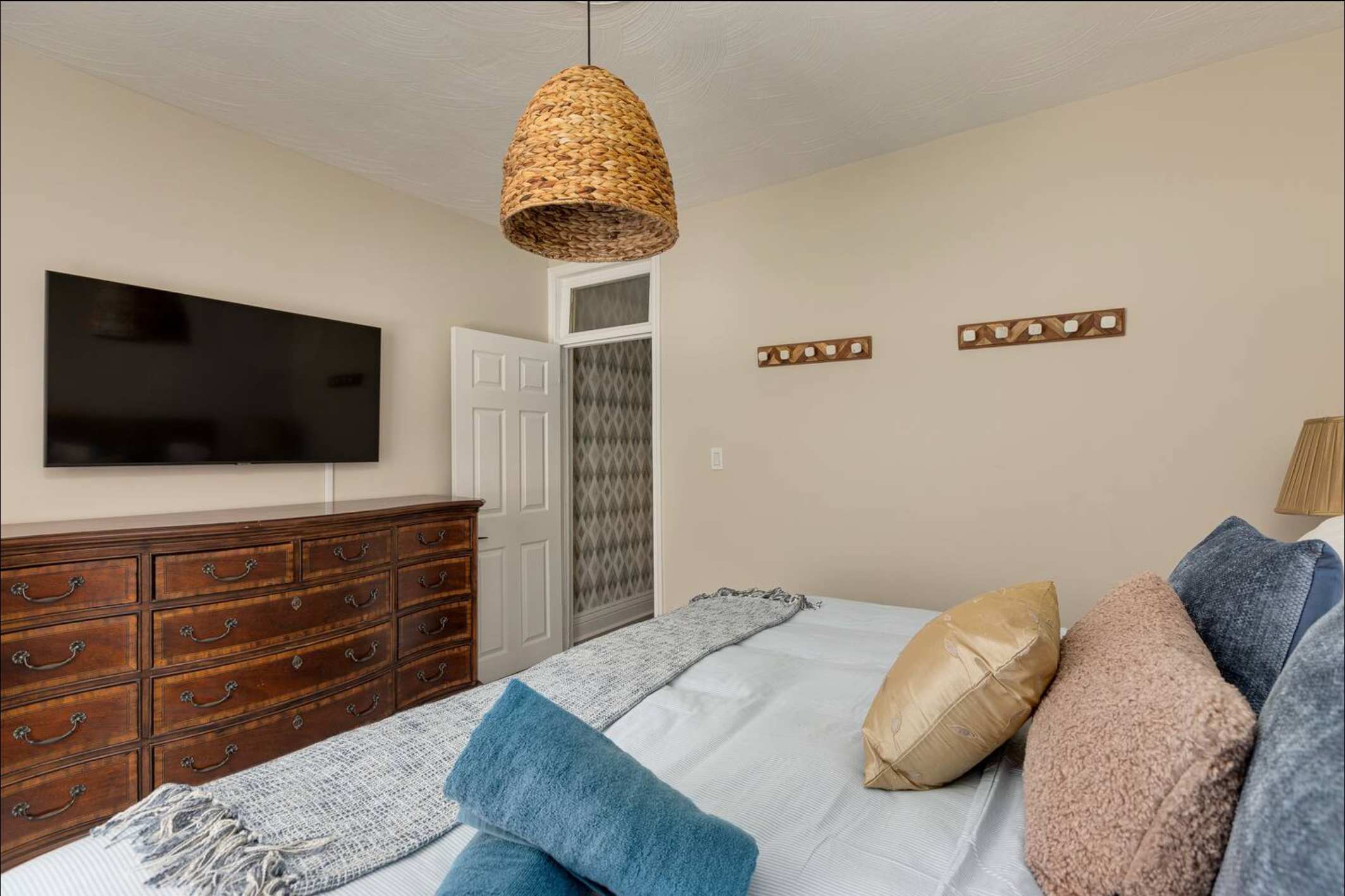 Cozy bedroom featuring a large dark wood dresser with multiple drawers, a wall-mounted flat-screen TV, and a bed with blue and gold pillows. A woven pendant light hangs from the ceiling.
