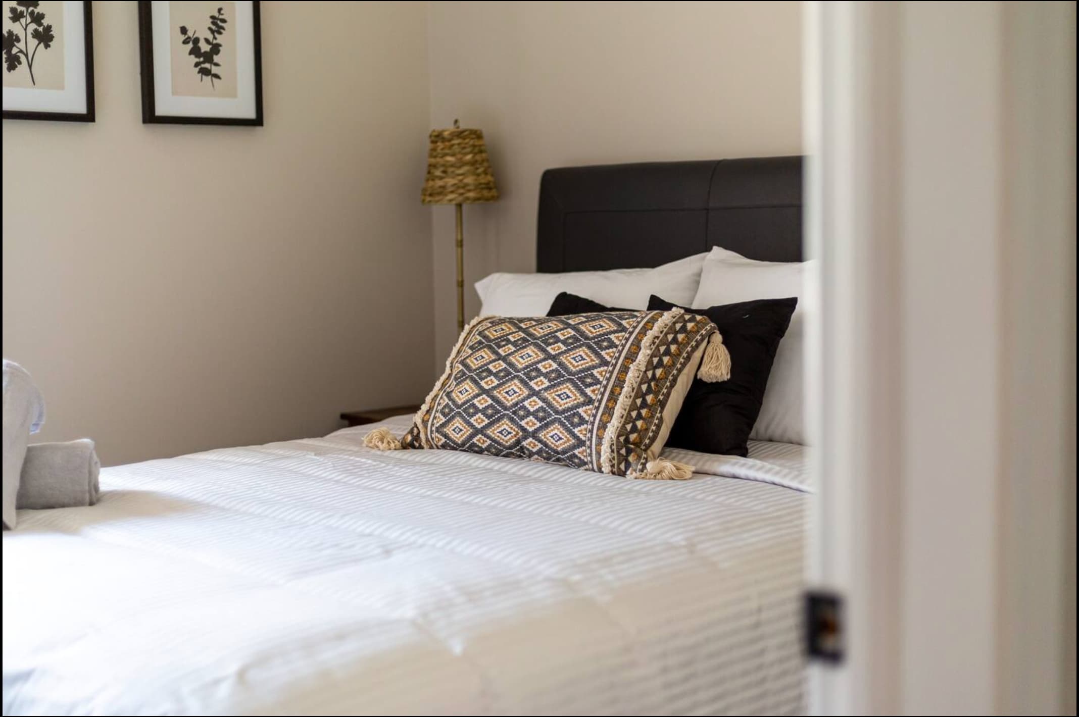 Close-up of a neatly made bed with white striped bedding, a patterned throw pillow, and two rolled grey towels. A dark upholstered headboard is visible behind the pillows.