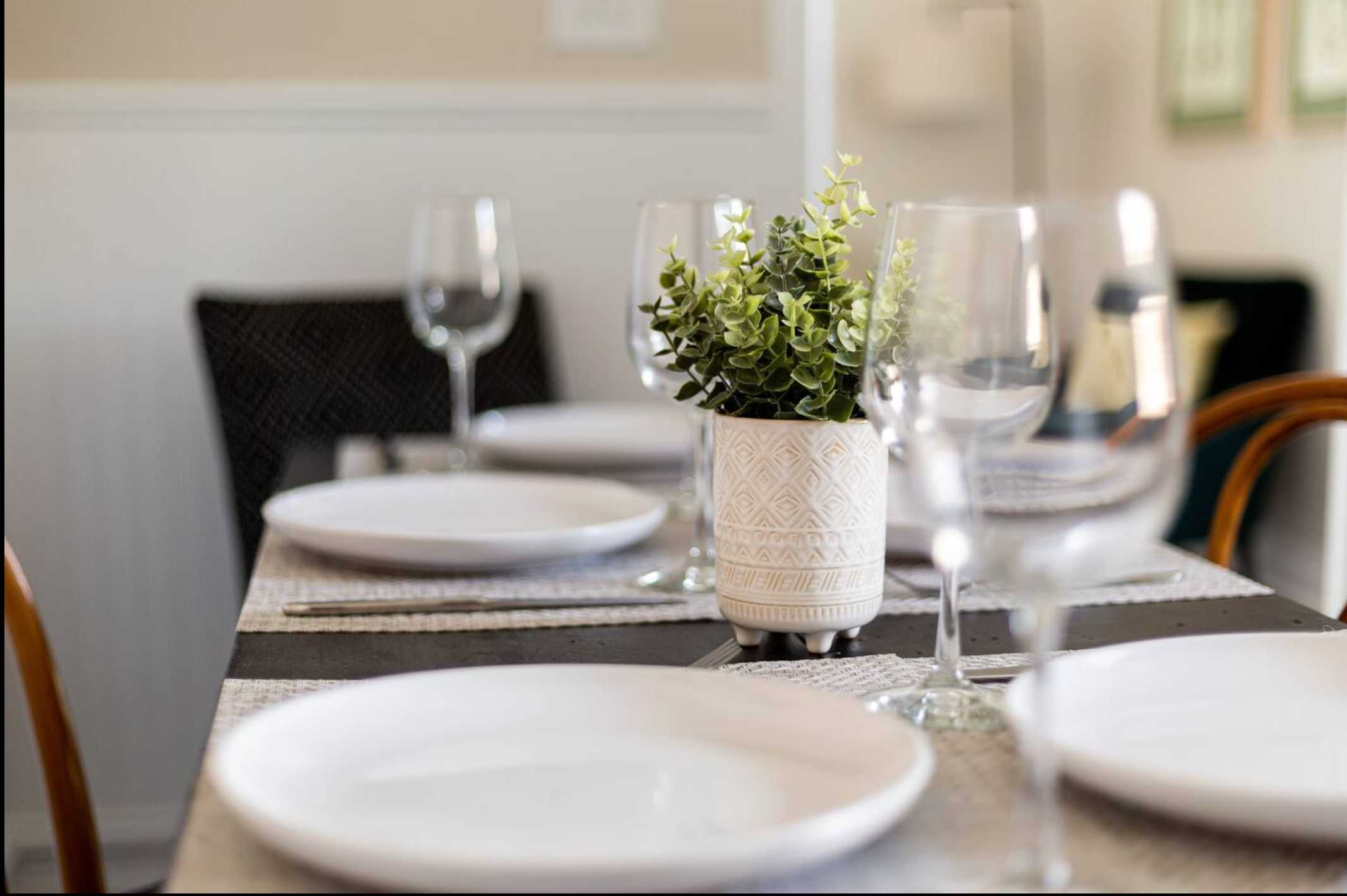 A close-up view of the dining table set with white plates, wine glasses, and placemats. A small potted green plant serves as a centerpiece, highlighting the table's dark surface.