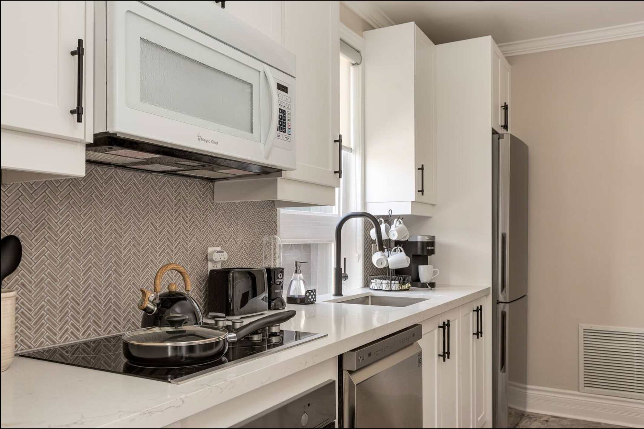 Bright kitchen featuring white shaker cabinets, a built-in microwave, an electric stovetop with a black kettle and pan, and a black gooseneck faucet over a stainless steel sink. A herringbone tile backsplash adds texture.