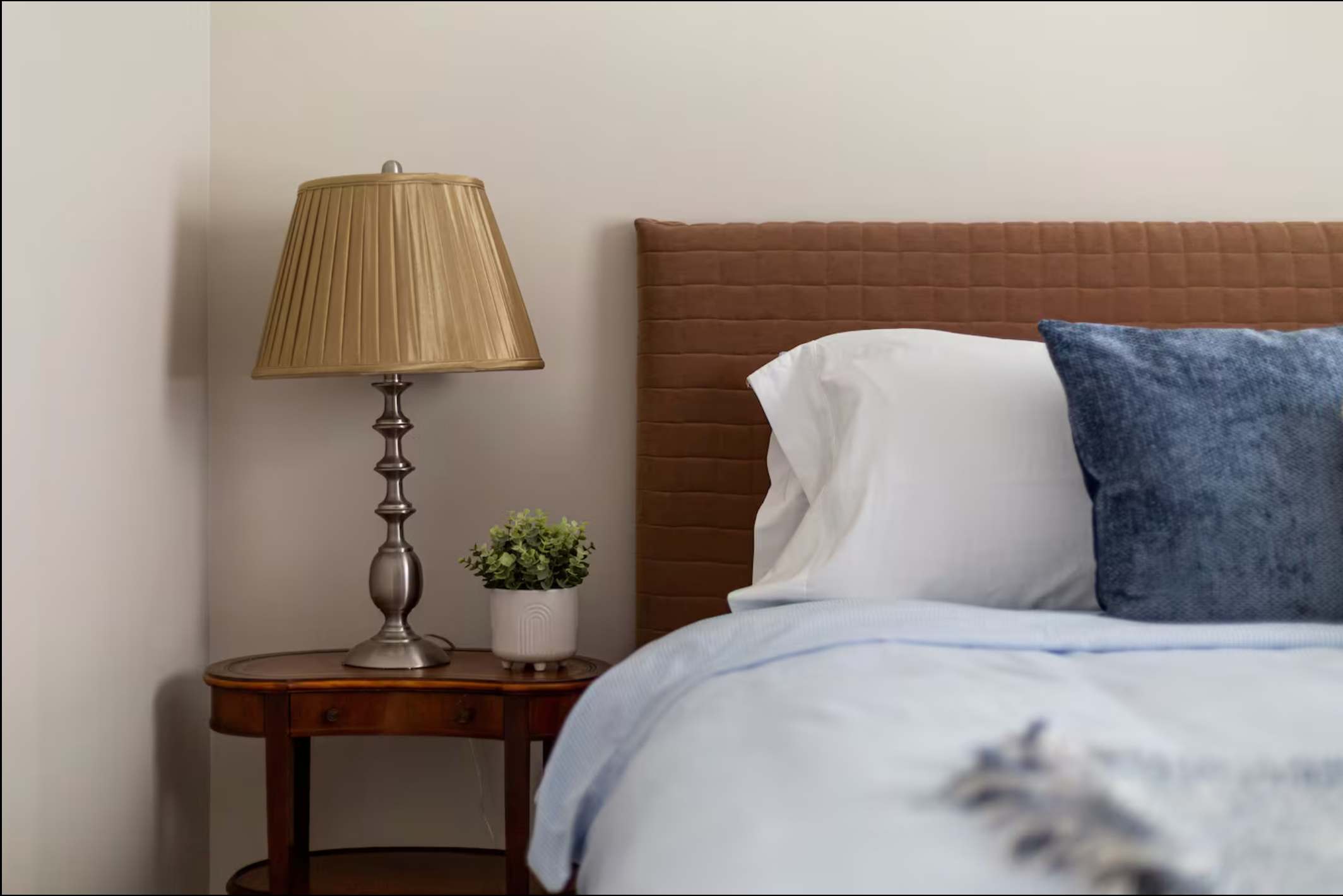 Close-up of a traditional wooden bedside table with a pleated gold lampshade and a small potted plant, next to a bed with a brown upholstered headboard and white and blue bedding.