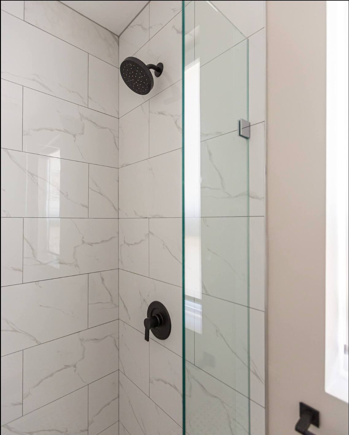 Modern shower with white marble-look subway tiles, a black rainfall showerhead, and a black single-handle mixer. A clear glass shower door separates the shower from the rest of the bathroom.