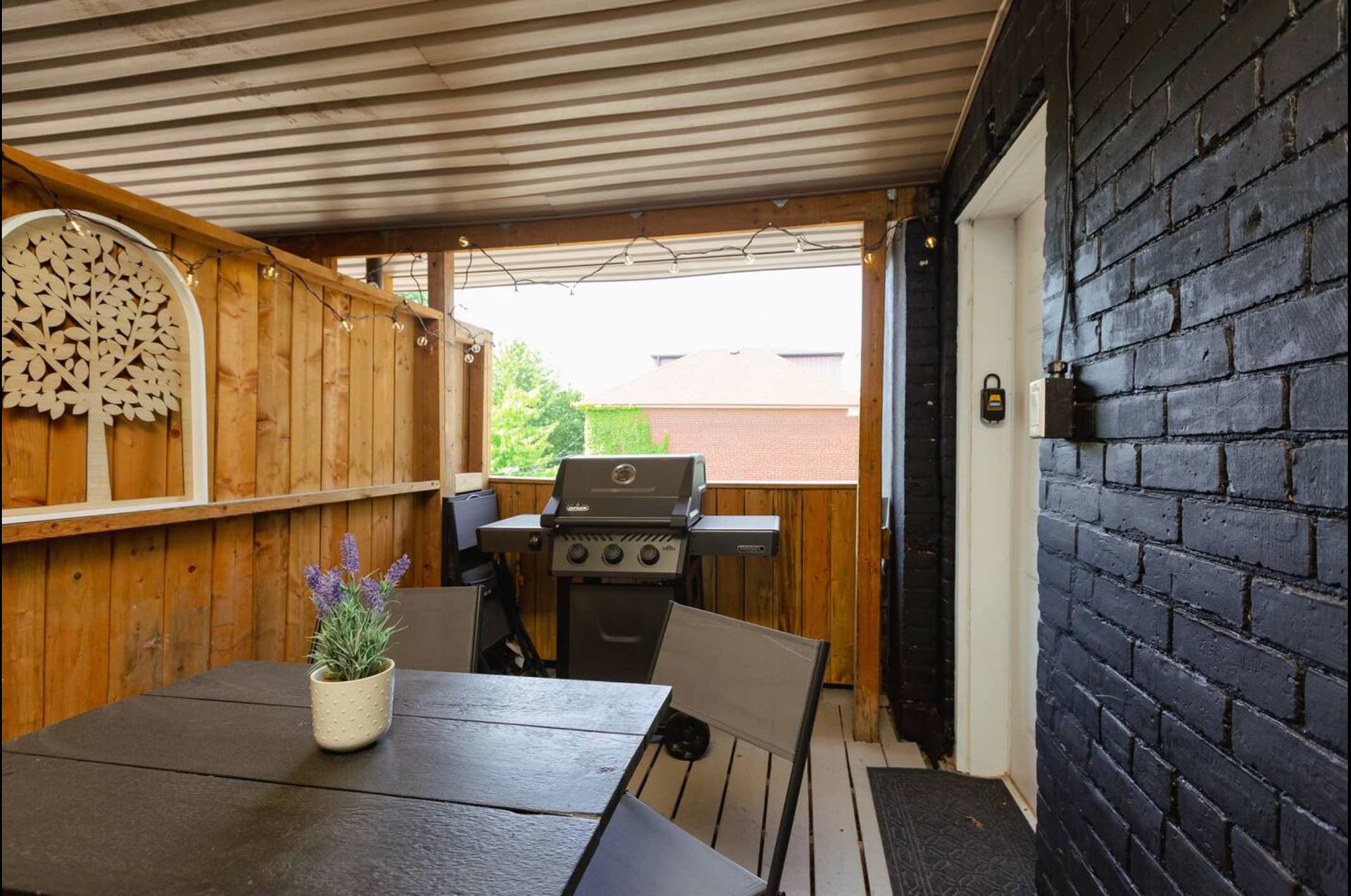 A covered outdoor patio features a dark grey dining set and a BBQ grill, enclosed by a dark brick wall and wooden fencing. String lights illuminate the space, offering a view of the neighborhood.