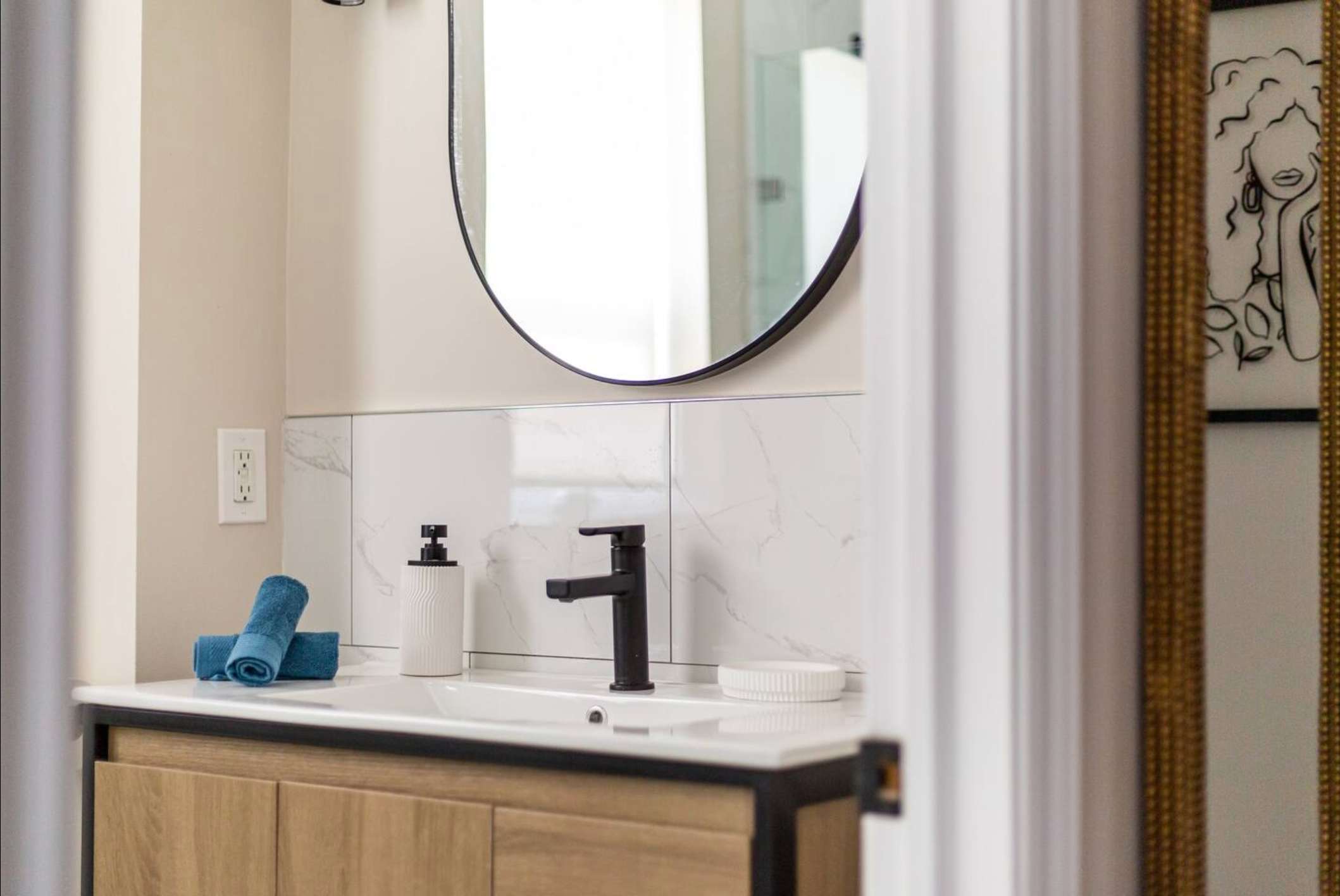 Close-up of a modern bathroom vanity with a light wood base, white countertop, and integrated sink. A black gooseneck faucet, soap dispenser, and round black-framed mirror are visible above white marble-look wall tiles.
