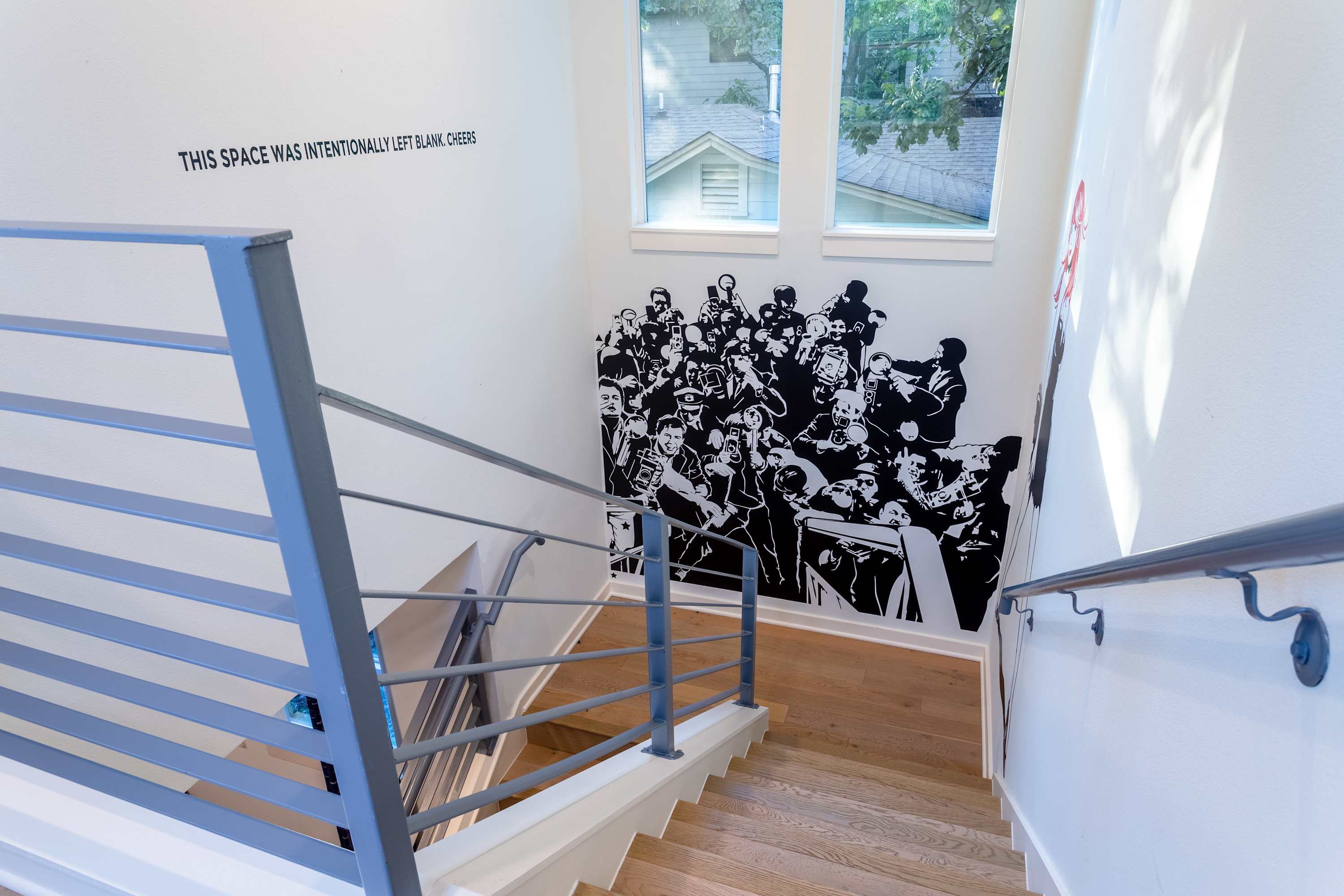 Modern staircase with light wood treads and gray metal railings, featuring a large black and white mural of a crowd on the wall and two windows above, providing natural light.