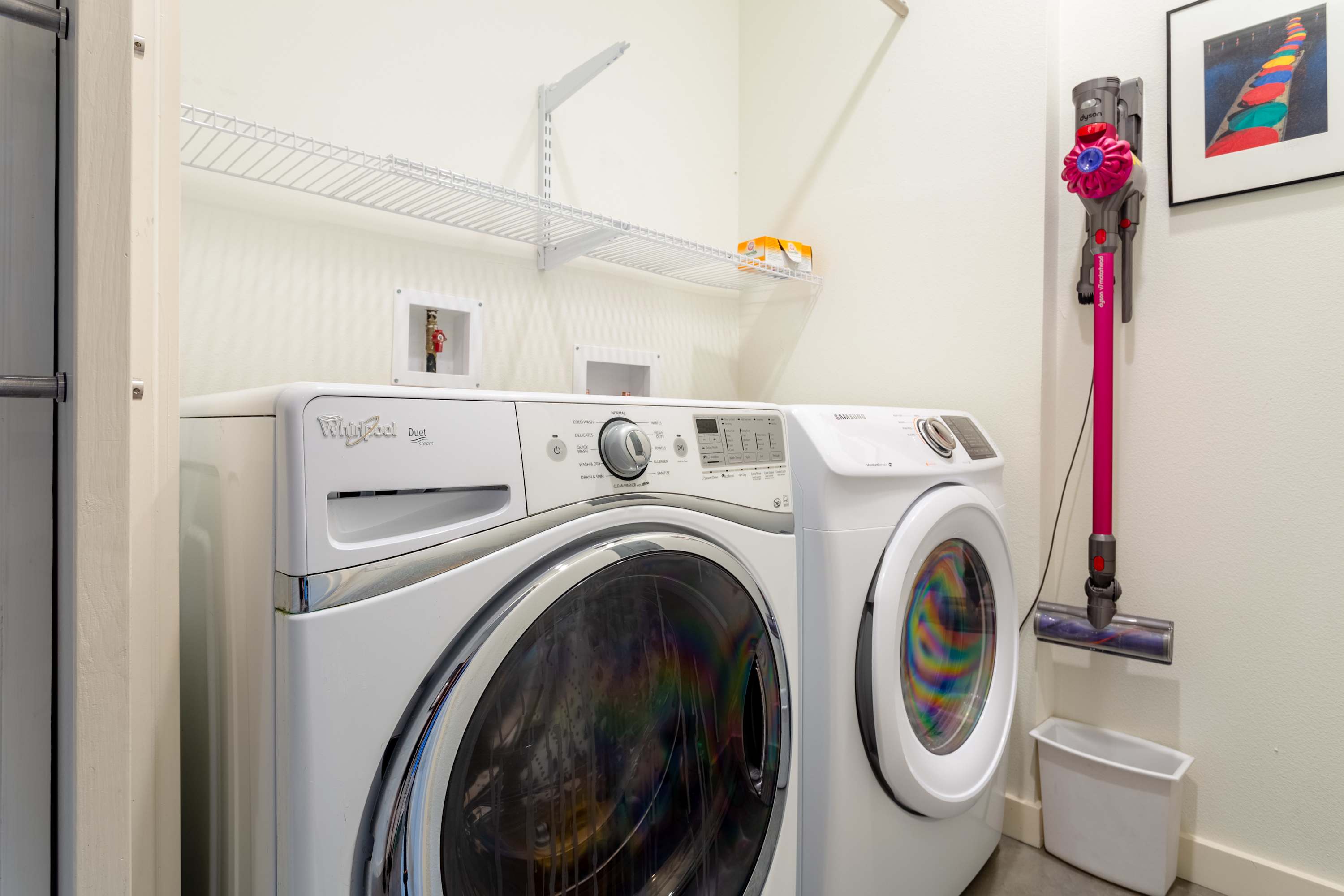 Functional laundry room equipped with a white Whirlpool washer and a Samsung dryer, overhead wire shelving, and a wall-mounted pink Dyson vacuum cleaner.