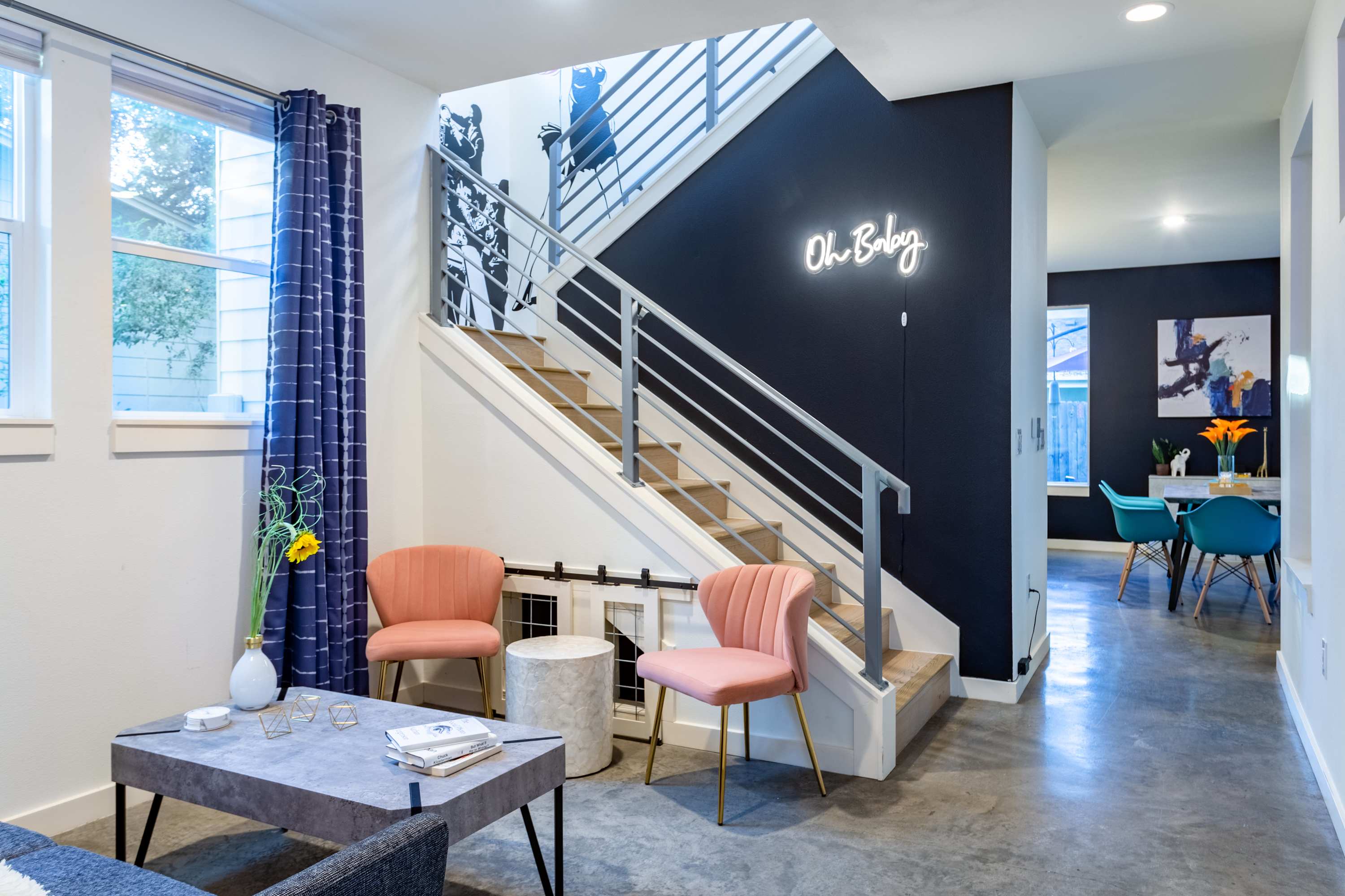 Modern living room with concrete floors, two pink velvet chairs, a grey coffee table, and a staircase with metal railings leading upstairs, featuring a "Oh Baby" neon sign on a dark accent wall.
