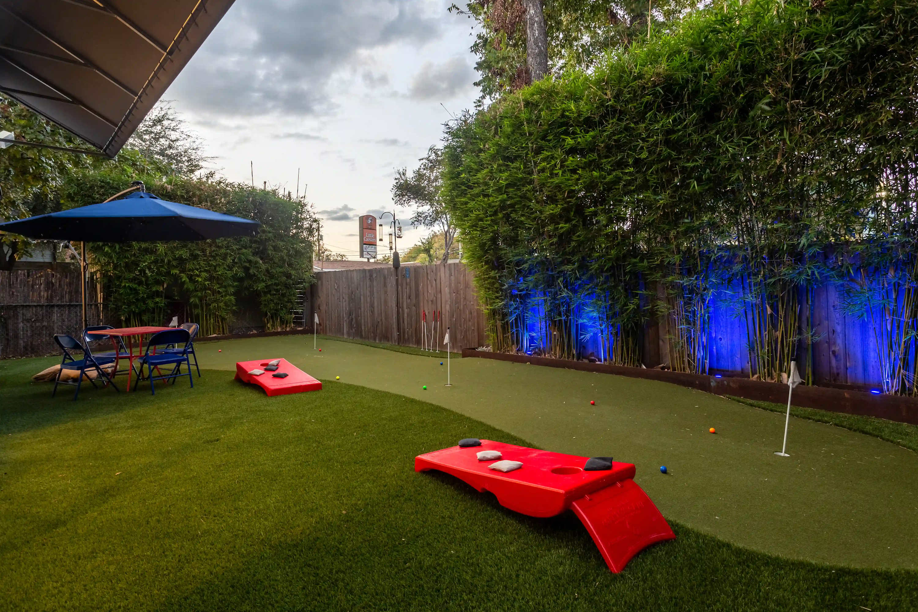 An expansive artificial turf backyard with a mini golf course and red cornhole boards, bordered by a tall bamboo hedge illuminated with blue accent lights. A blue umbrella shades a small seating area.