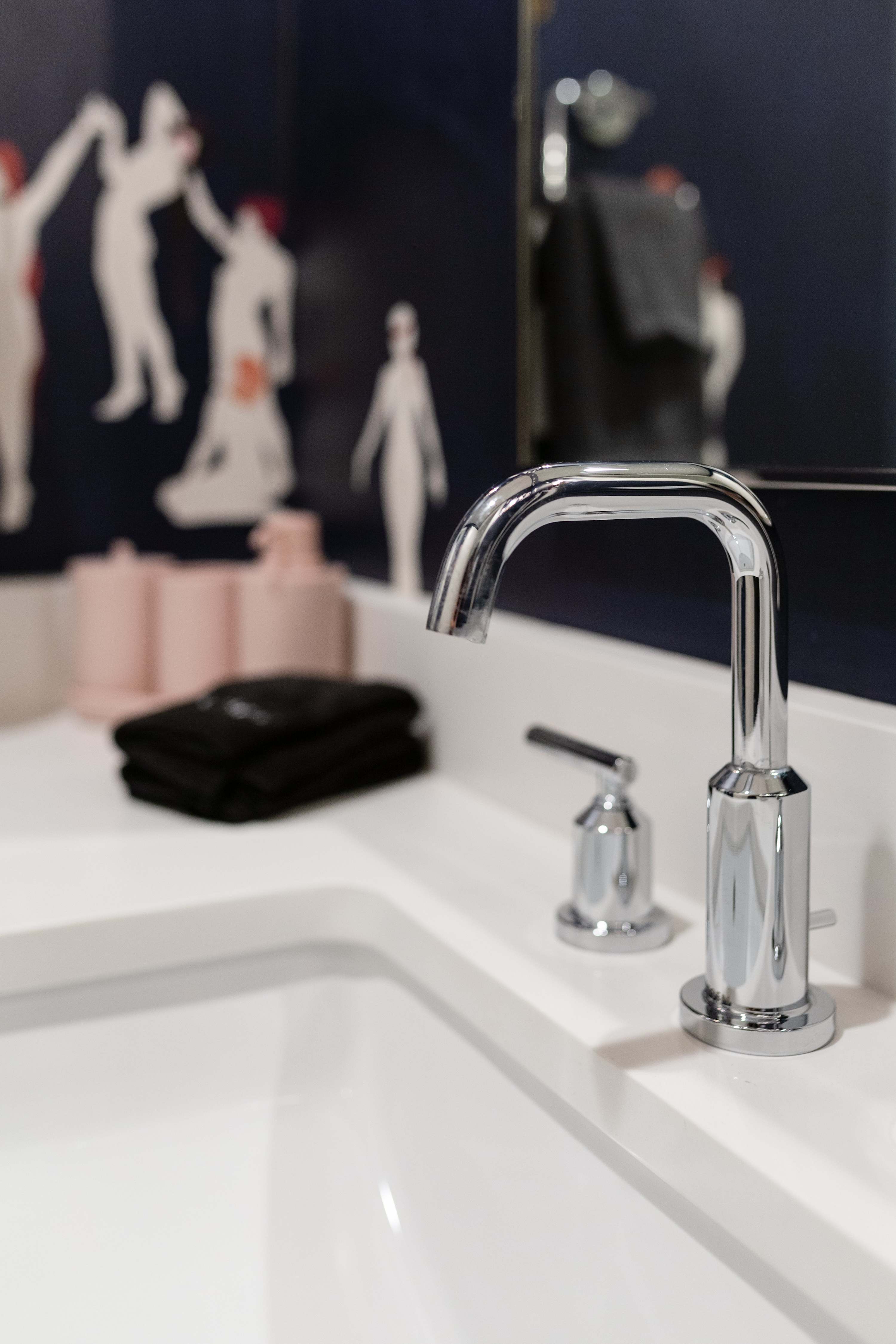 Close-up of a modern chrome gooseneck faucet and sink on a white quartz countertop, with a dark blue wall featuring white and red abstract figures in the background.