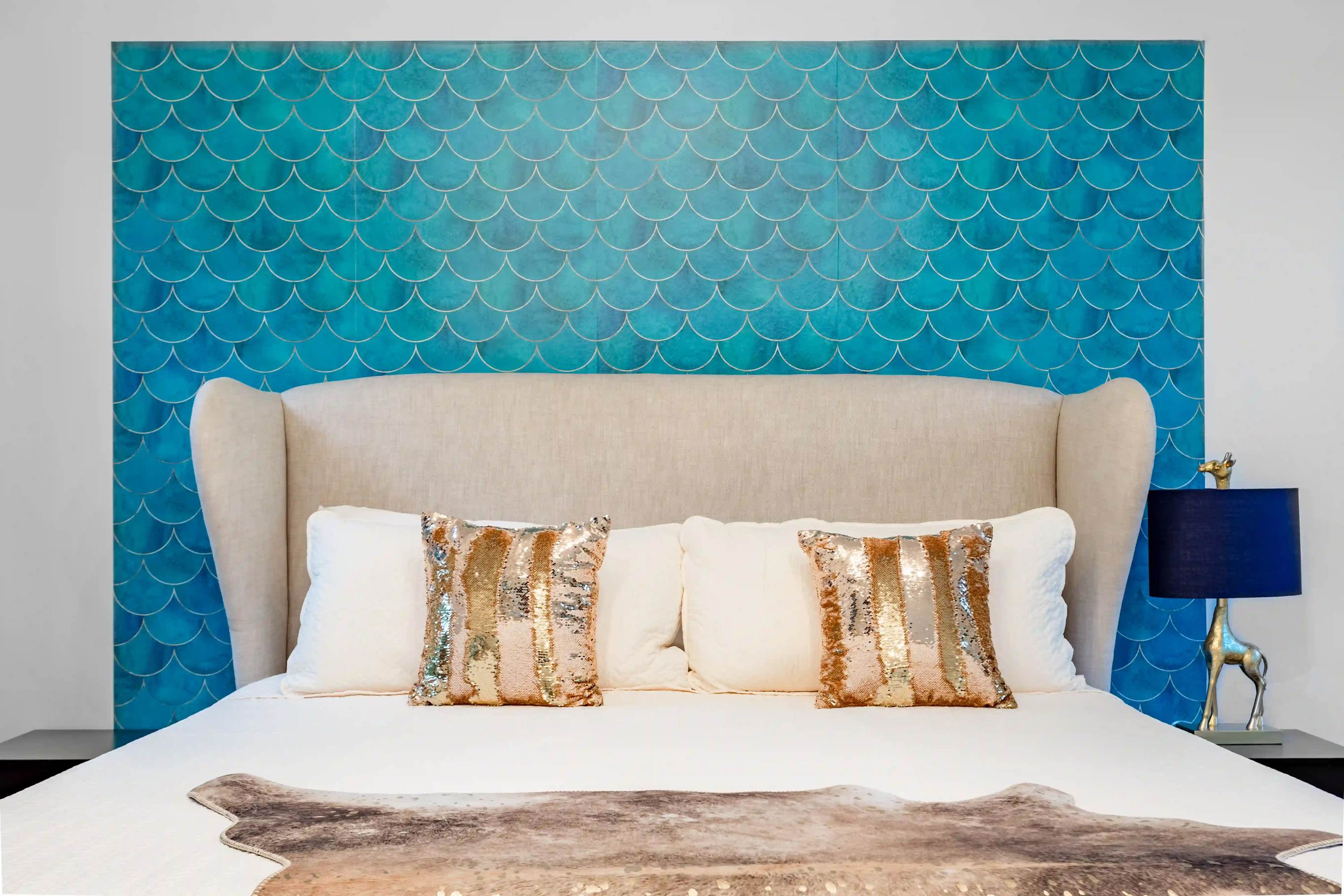 Bedroom featuring a large bed with a tall upholstered headboard, two sequined throw pillows, and a striking teal blue scalloped pattern accent wall behind the bed.