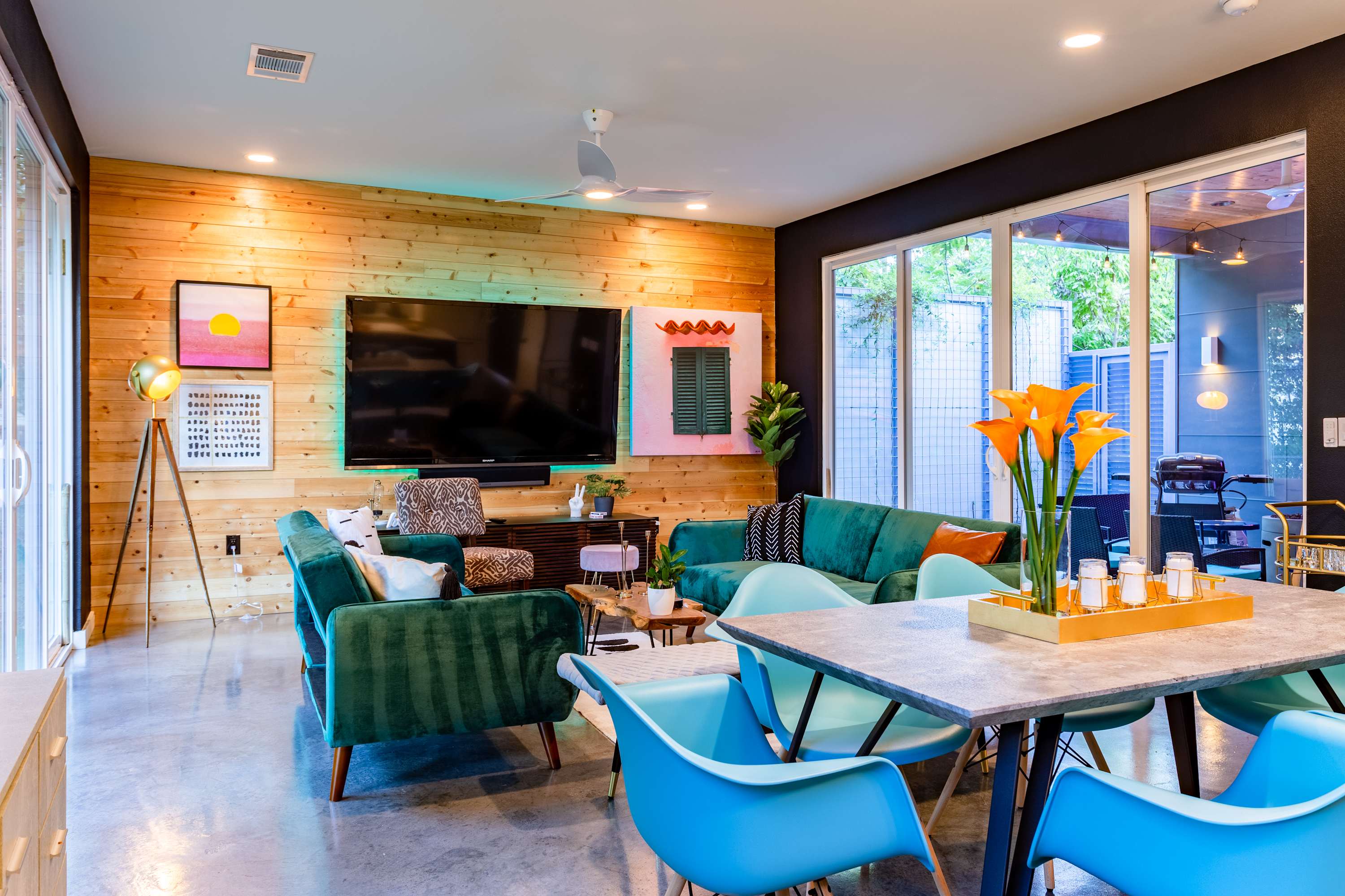 A spacious living room with two teal velvet sofas, a large flat-screen TV on a wood plank accent wall, and concrete floors. Large sliding glass doors connect to an outdoor patio, and a dining area is visible in the background.