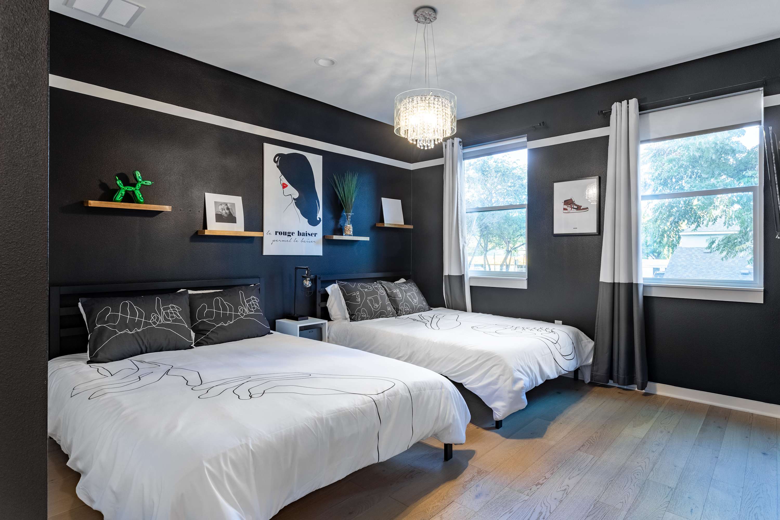 Stylish bedroom with two beds featuring white bedding with black line art, dark gray walls, a crystal chandelier, and two large windows with gray and white curtains.