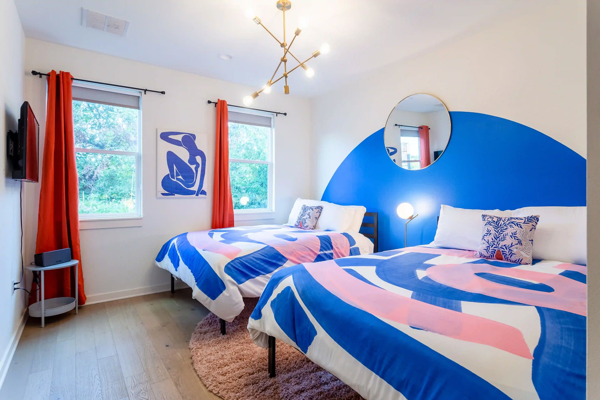 Vibrant bedroom with two beds featuring bold blue and pink abstract bedding, a large blue arched accent wall, a modern chandelier, and two windows with orange curtains.