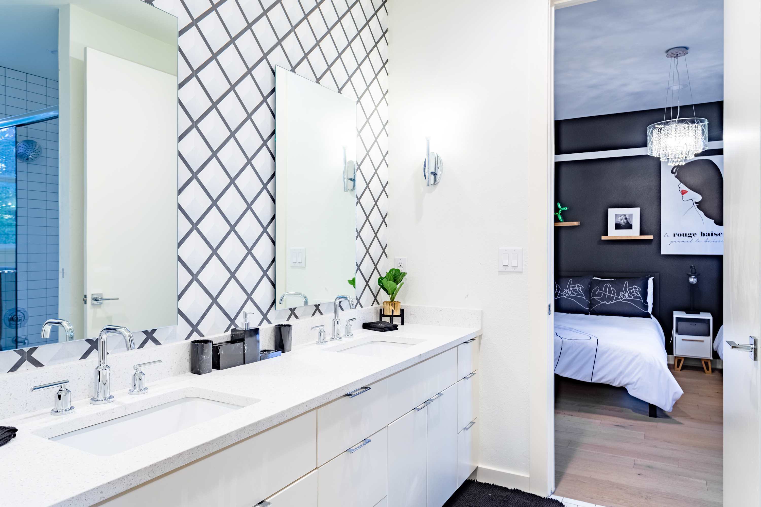 Modern bathroom with a double vanity, white quartz countertops, chrome faucets, and a striking black and white geometric patterned accent wall behind the mirrors, with a view into a dark-walled bedroom.