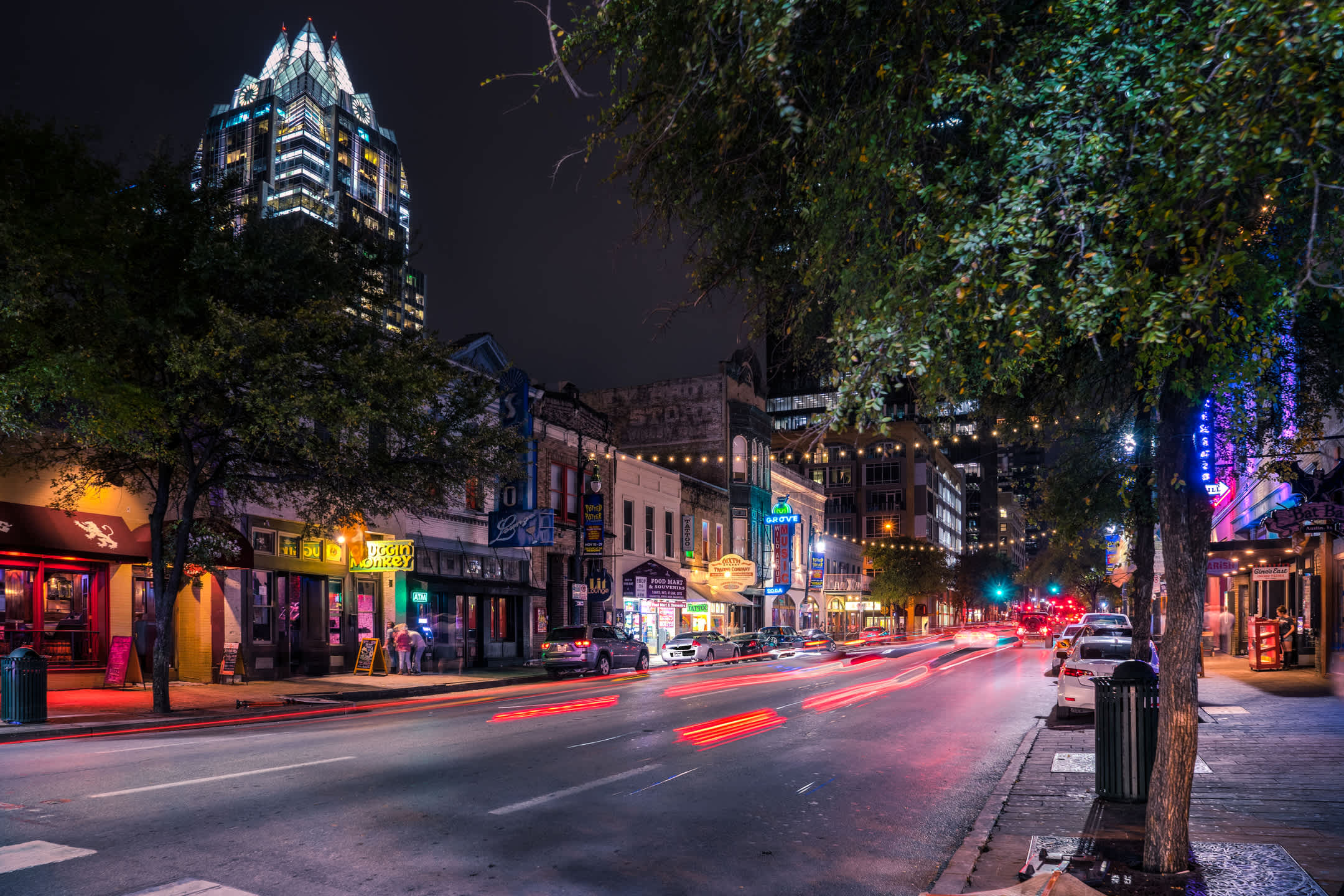 Dynamic night view of a bustling downtown street with blurred red car lights, illuminated storefronts, and a tall, modern skyscraper in the background under a dark sky.