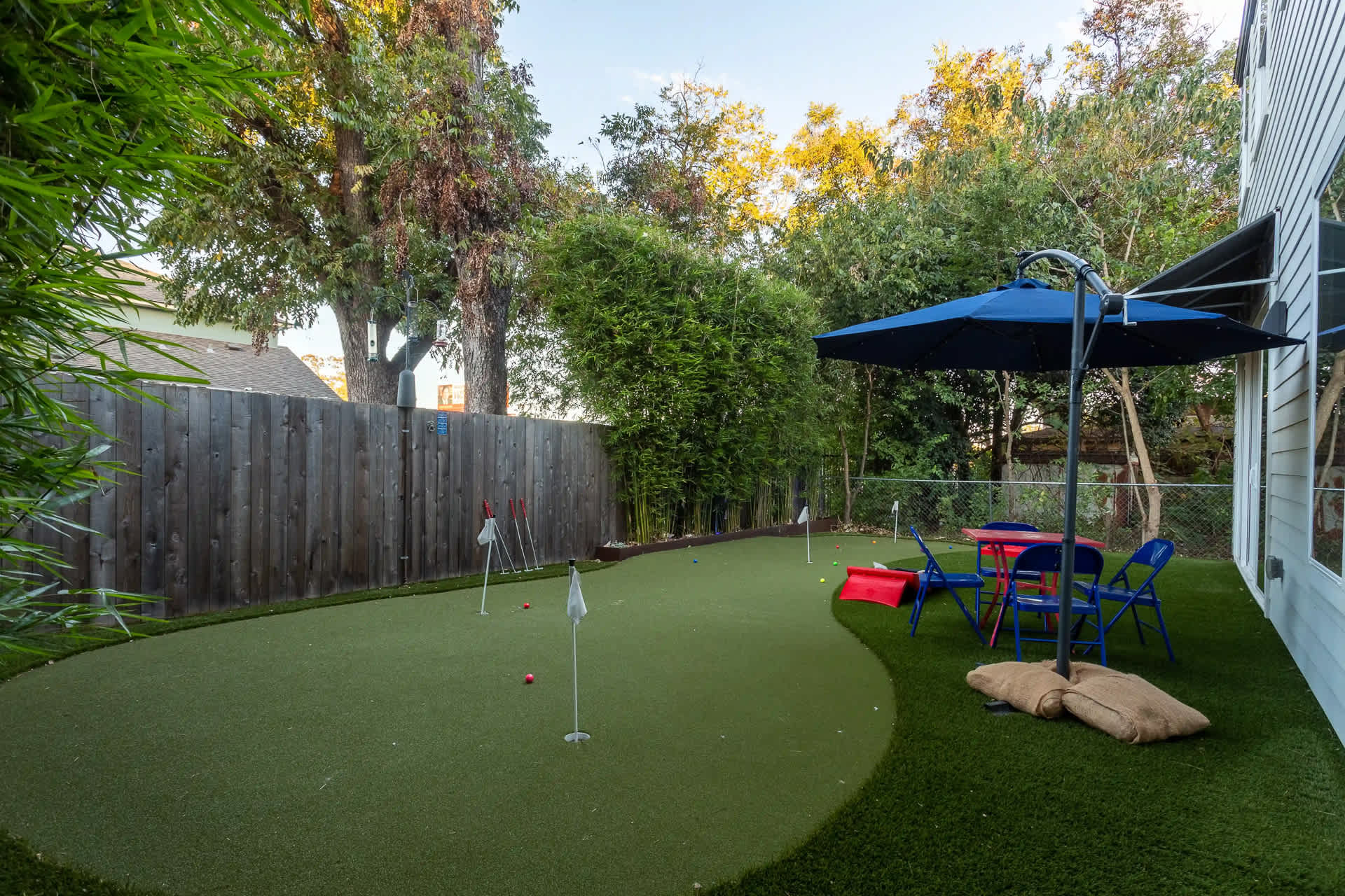 A vibrant backyard features an artificial turf putting green with multiple holes and flags, alongside a small red table and blue chairs under a blue umbrella. Tall bamboo plants line the fence, creating a private oasis.