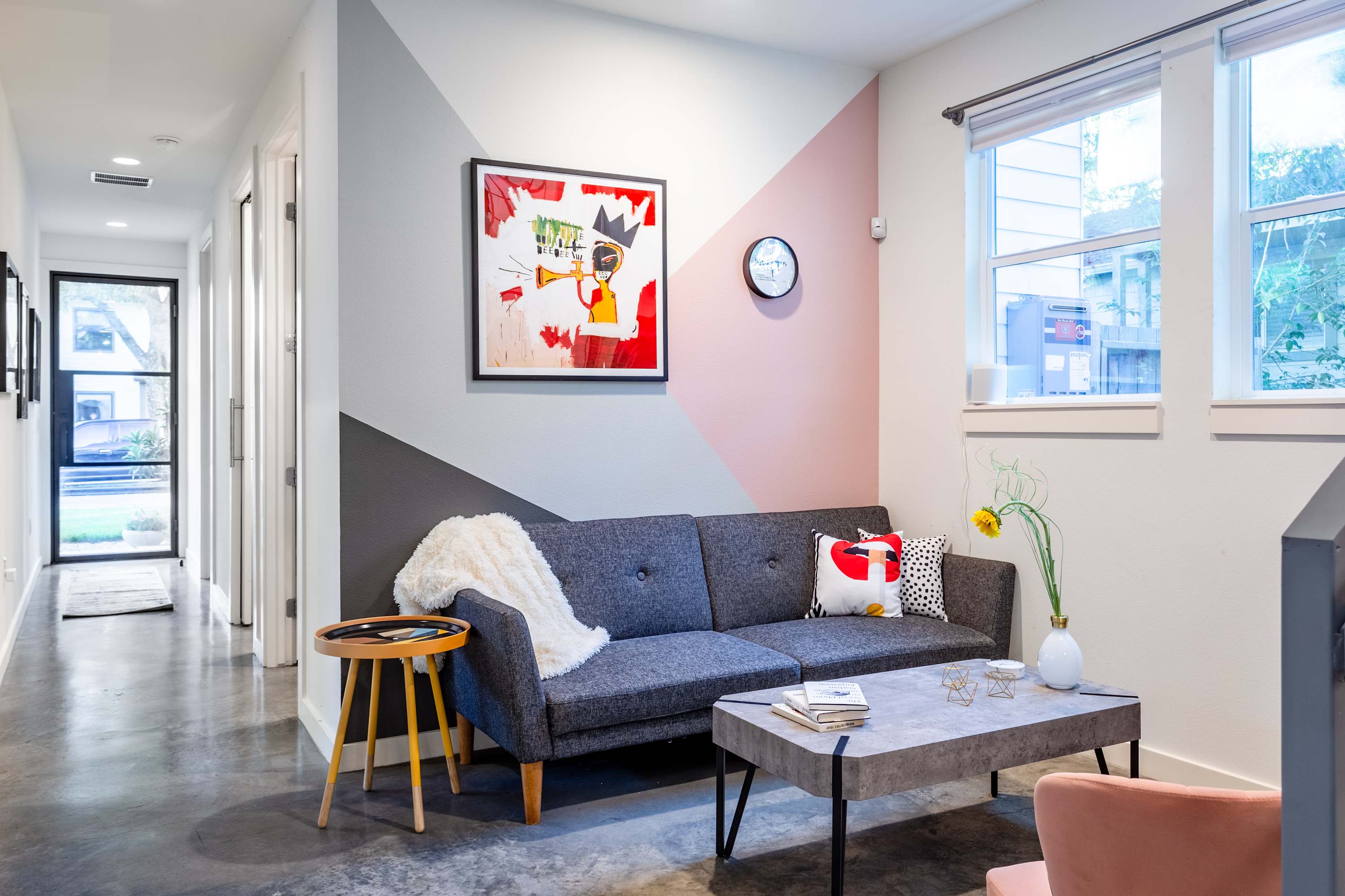 Cozy living area with a grey sofa, a geometric accent wall painted in grey, pink, and white, a modern coffee table, and a small wooden side table, illuminated by natural light from a window.