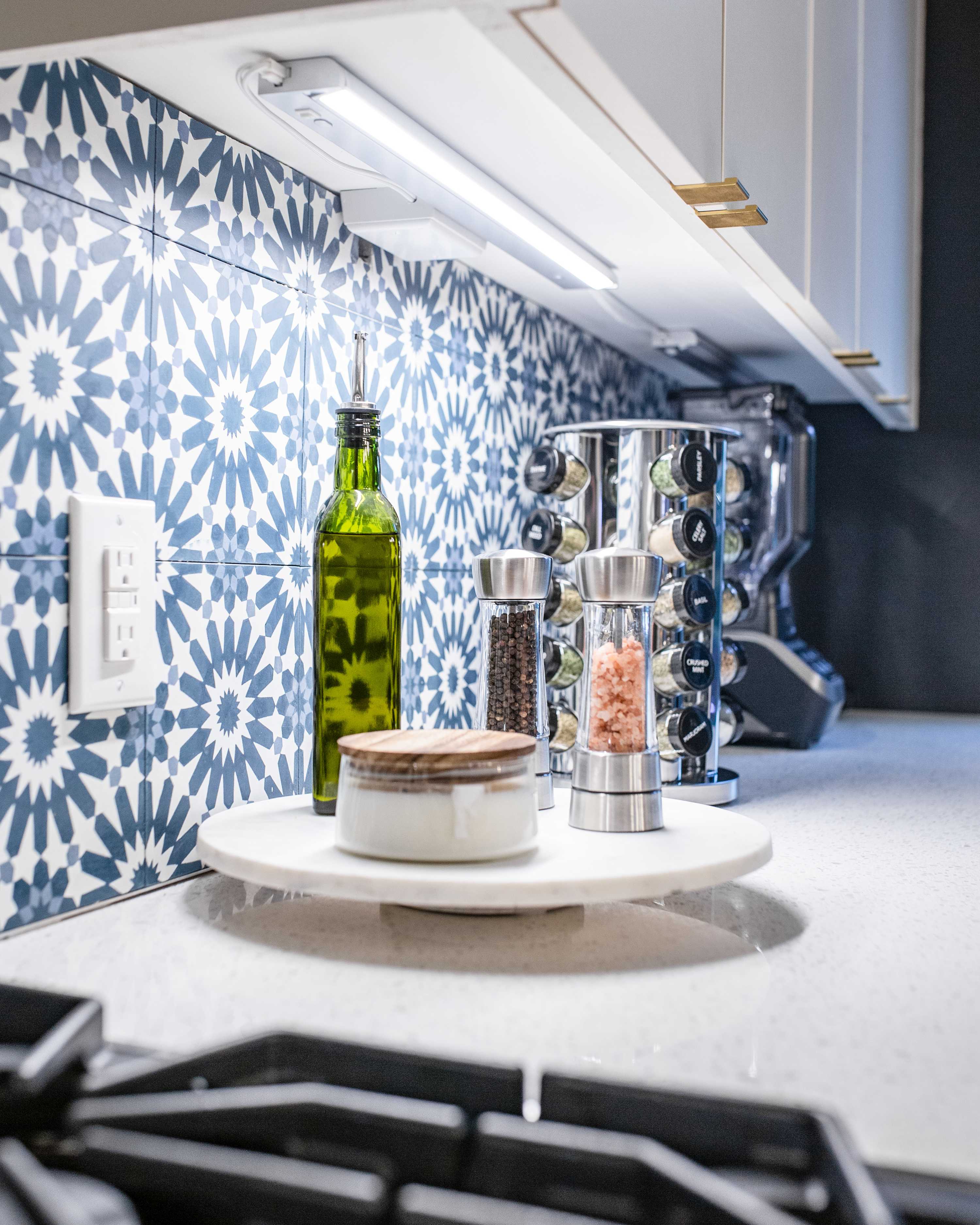 Close-up of a kitchen counter featuring a white marble lazy Susan holding an olive oil bottle, salt and pepper grinders, and a small container, set against a blue and white patterned tile backsplash.