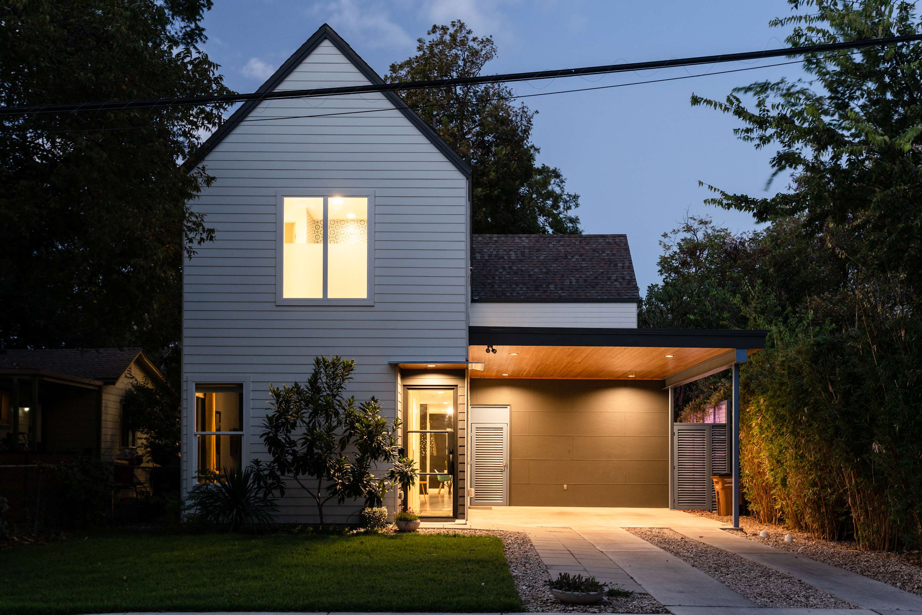 The modern two-story house exterior at dusk, featuring light-colored siding, a dark roof, and a covered carport. Warm lights illuminate the entrance and carport, highlighting the contemporary design.