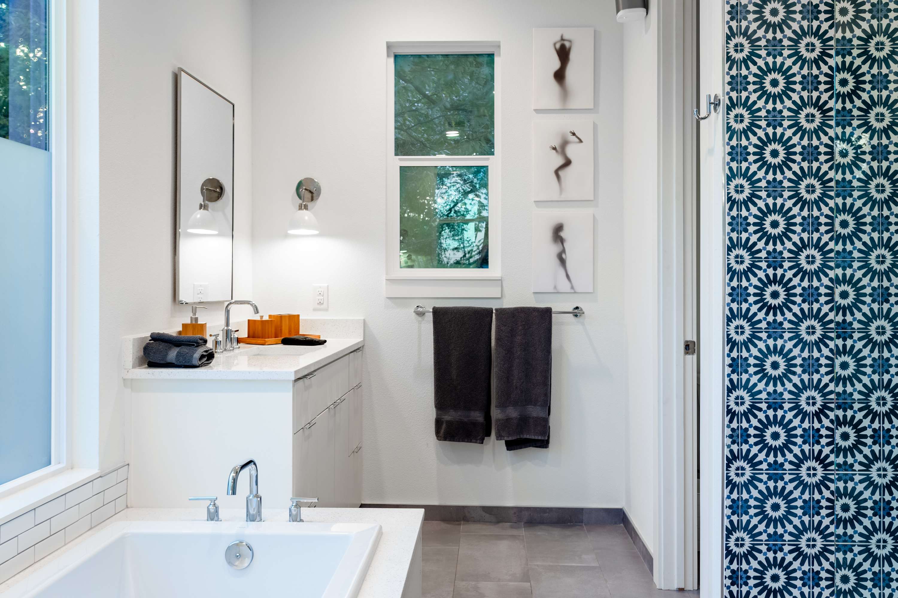 Modern bathroom with a white vanity, a large mirror, and a window, featuring a striking blue and white patterned tile wall near the doorway, and three framed artworks above the towel rack.