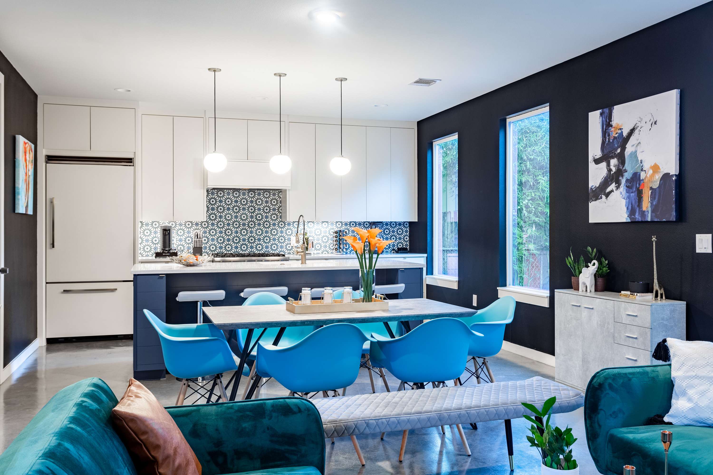 An open-concept dining area with a long gray table and six bright blue chairs, adjacent to a modern kitchen with white cabinets and a patterned blue and white backsplash. Dark accent walls provide a striking contrast.