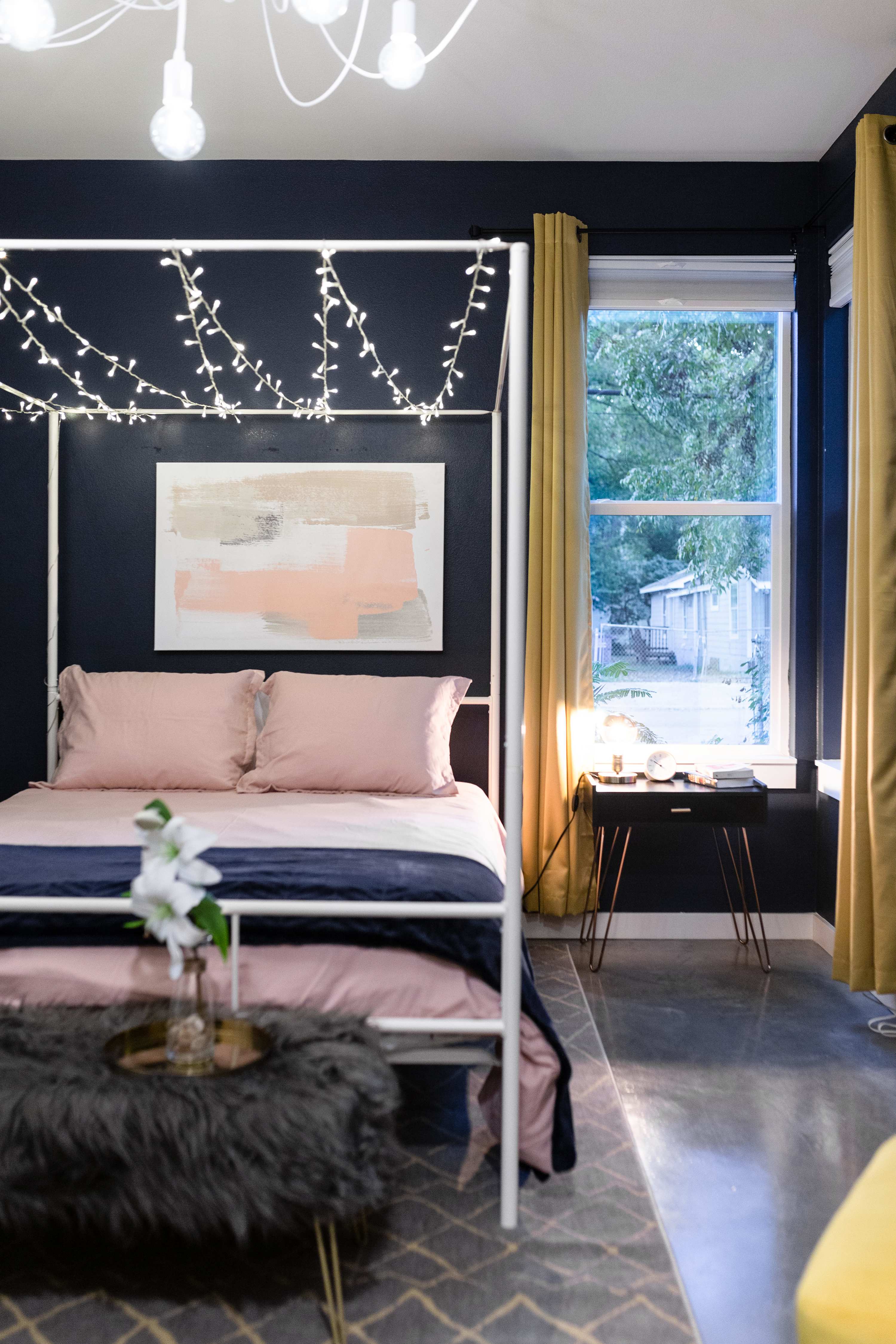 Close-up view of a dark blue bedroom's canopy bed with string lights, pink bedding, and a modern black nightstand next to a window with yellow curtains, offering a glimpse of the outdoors.