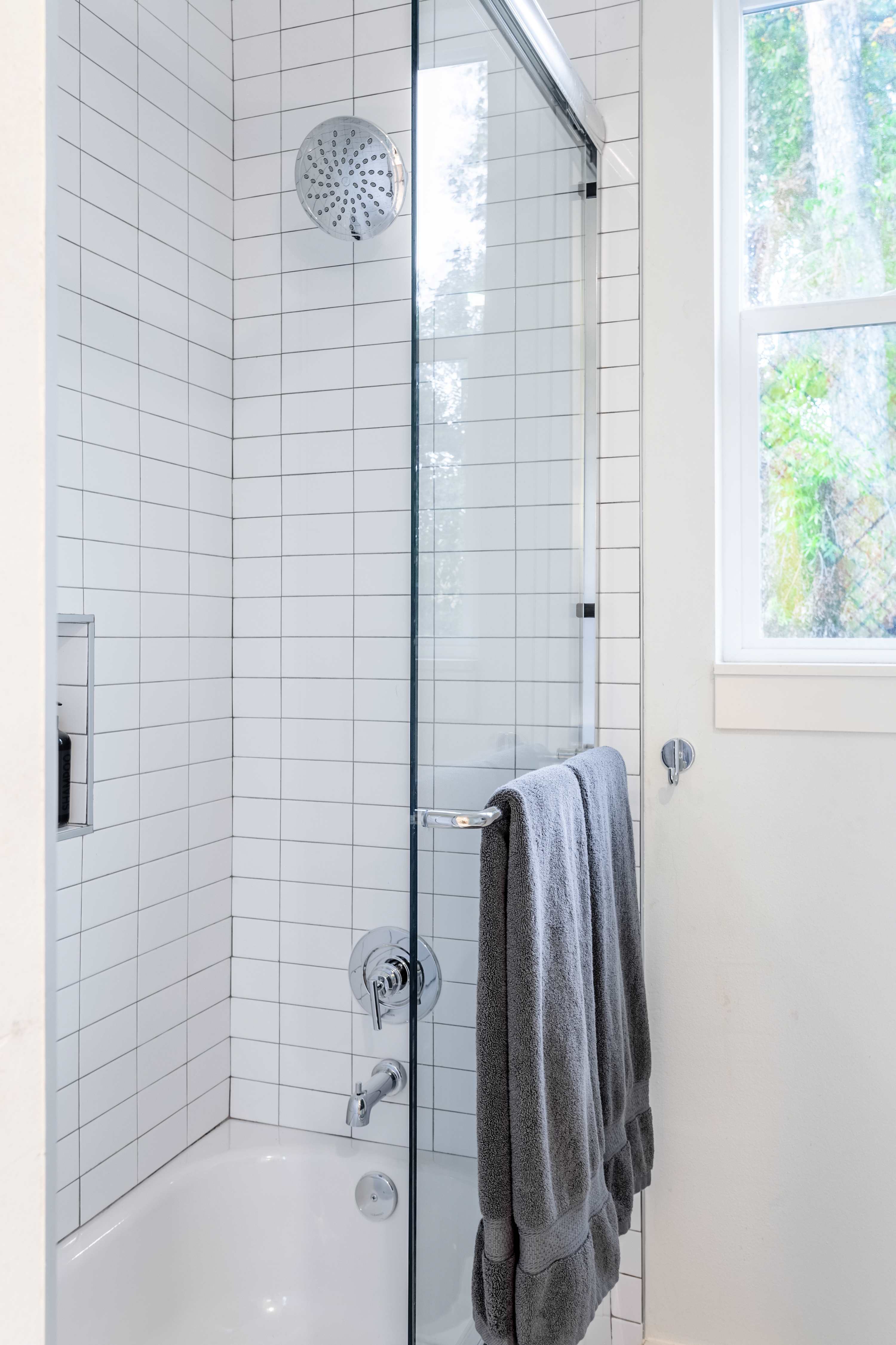 Bright bathroom featuring a white tiled shower/tub combo with a glass enclosure, a chrome showerhead, and a window providing natural light.