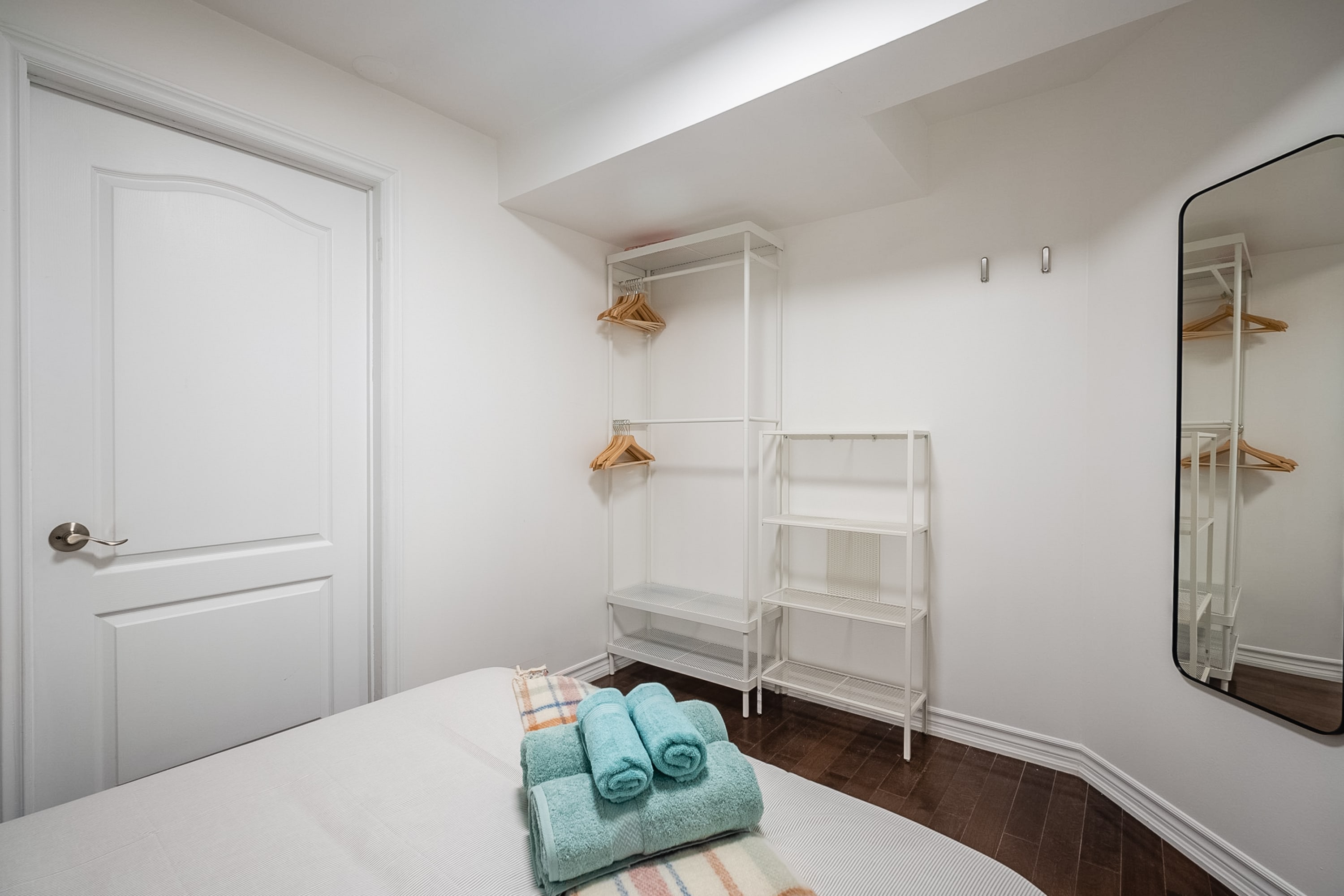A different perspective of the bedroom, showcasing a white paneled door and a white open closet system with wooden hangers. A full-length mirror reflects the closet, and the bed with blue towels is partially visible.