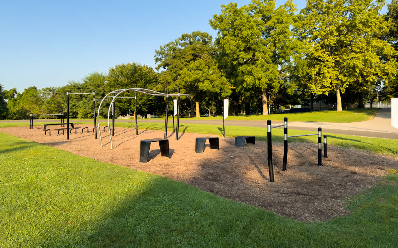 An outdoor fitness park with various metal exercise structures, including pull-up bars and benches, set on a wood chip ground. The area is surrounded by lush green grass and mature trees under a clear blue sky.