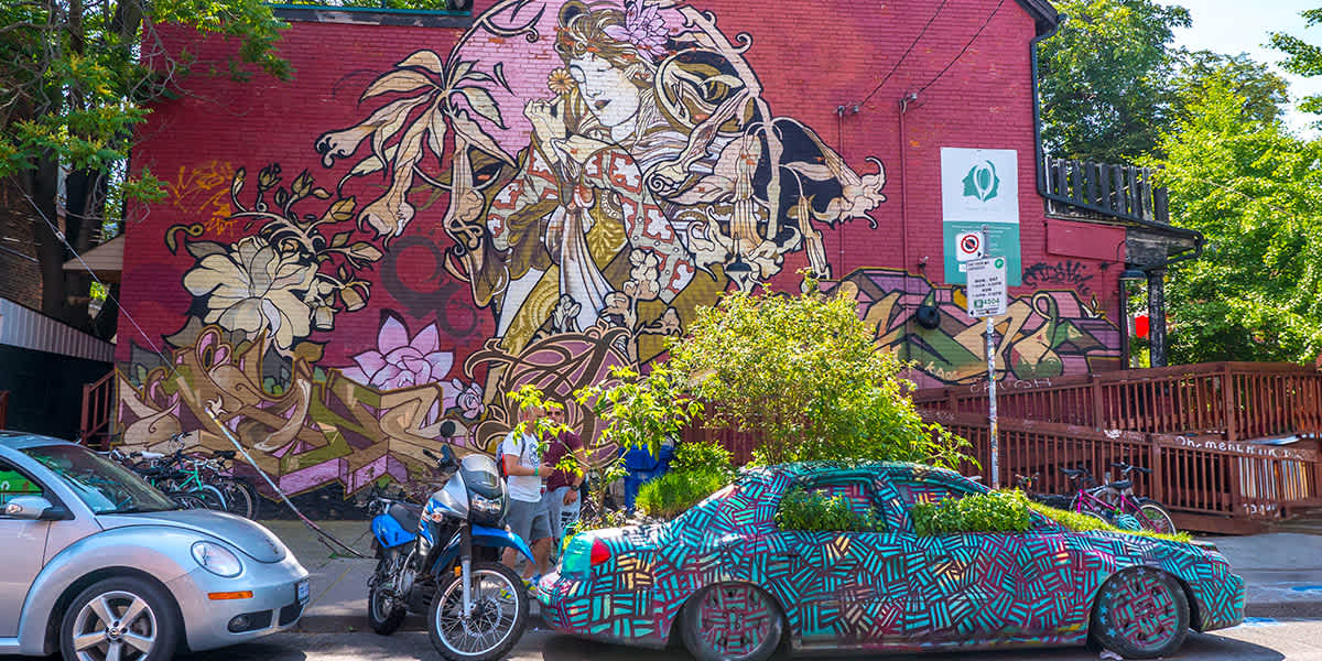 A vibrant urban scene featuring a large, colorful mural on a red brick building, depicting a woman with flowing hair and floral patterns. A uniquely decorated car with plants growing from it is parked below, alongside other vehicles.