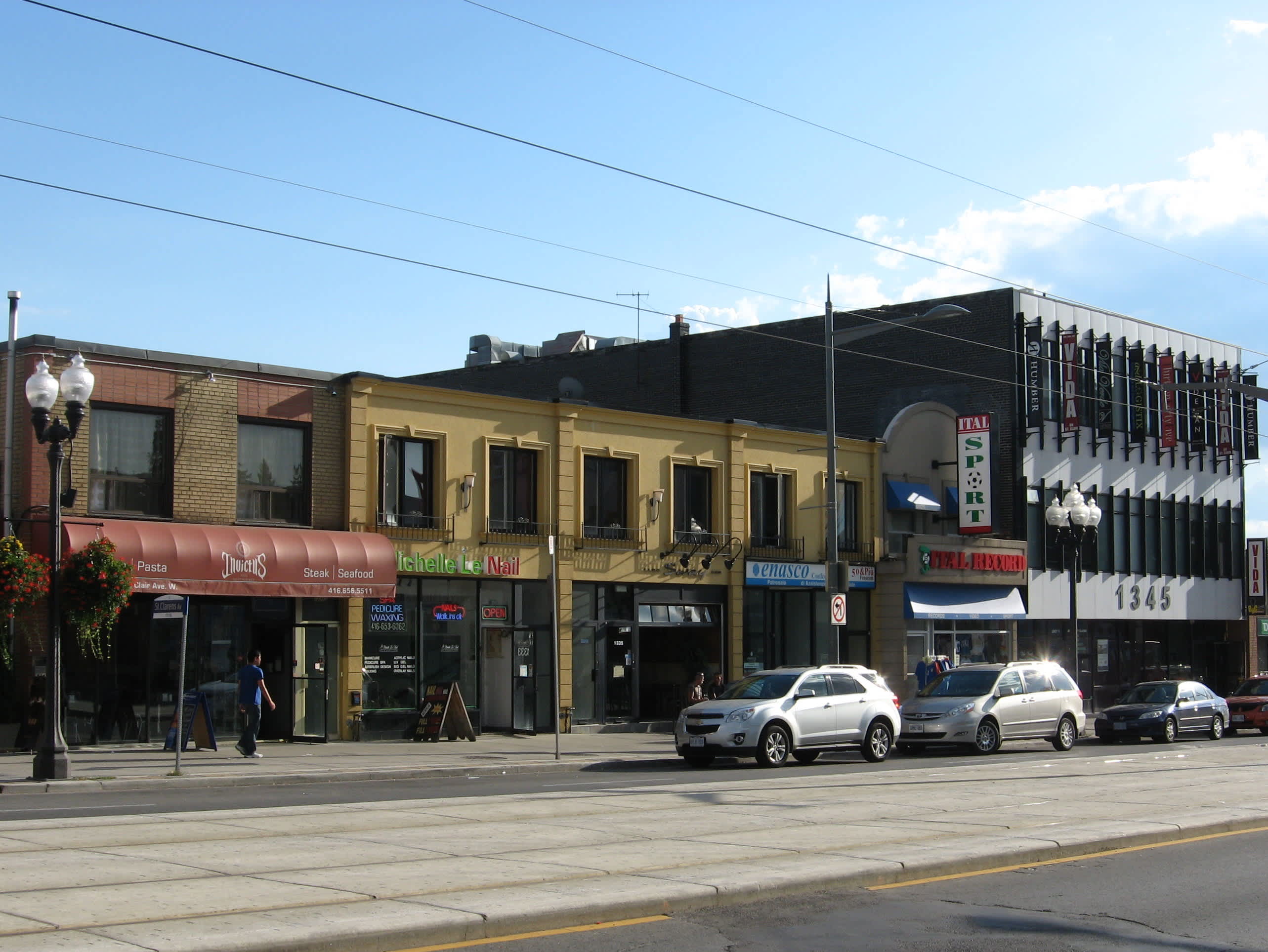A bustling street scene showcasing a row of diverse commercial buildings with varying architectural styles, from brick facades to modern structures. Parked cars line the street, and a streetcar track is visible in the foreground.