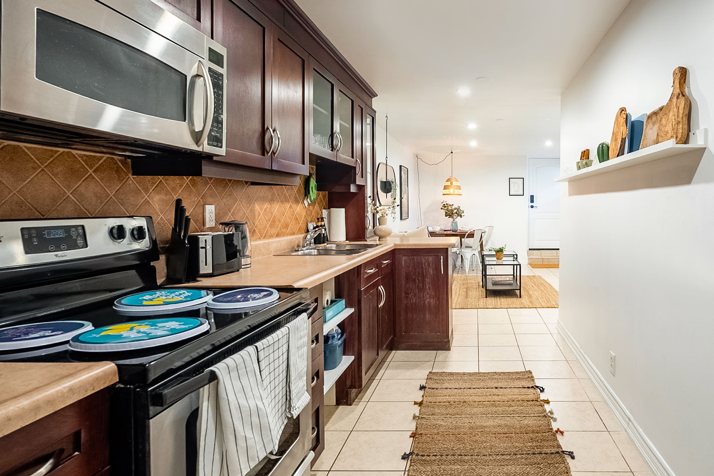 A well-equipped kitchen features dark cherry wood cabinets, light countertops, and a tiled backsplash, with stainless steel appliances including a microwave and stove. A small jute rug lies on the tiled floor, and a white floating shelf with decorative items is on the opposite wall.