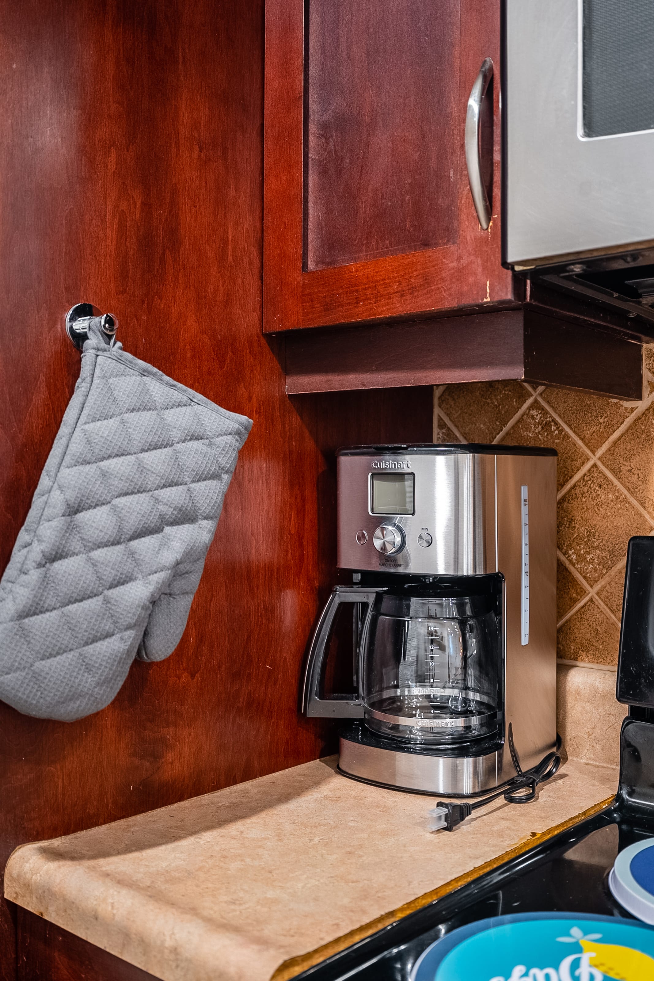 A close-up shows a silver Cuisinart coffee maker on a light beige countertop in the kitchen, next to dark cherry wood cabinets. A gray oven mitt hangs on the cabinet door, indicating a functional cooking space.
