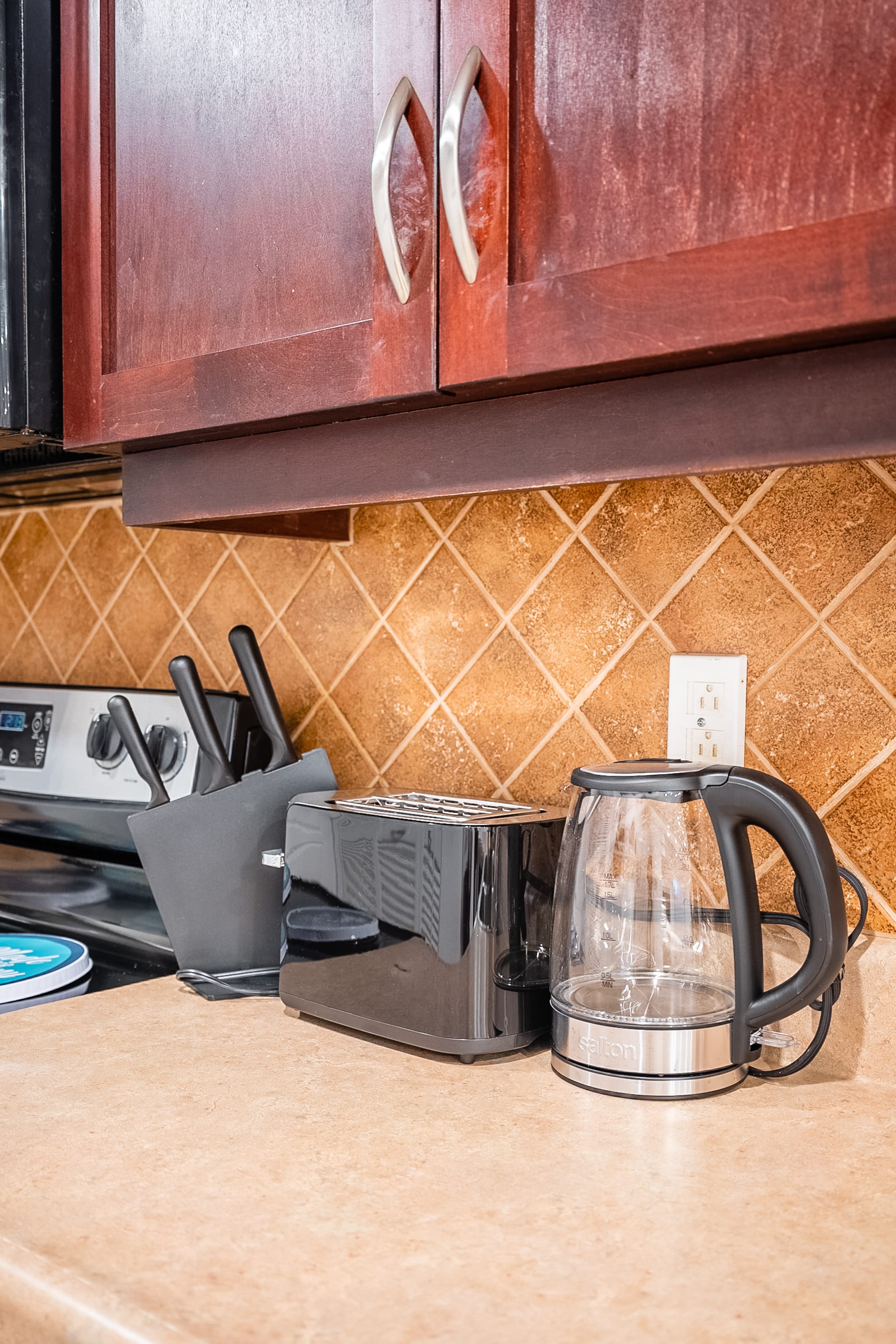 A close-up view of a kitchen counter featuring a sleek black toaster, a clear electric kettle, and a black knife block. The backsplash is adorned with small square brown tiles, complemented by dark wood upper cabinets.