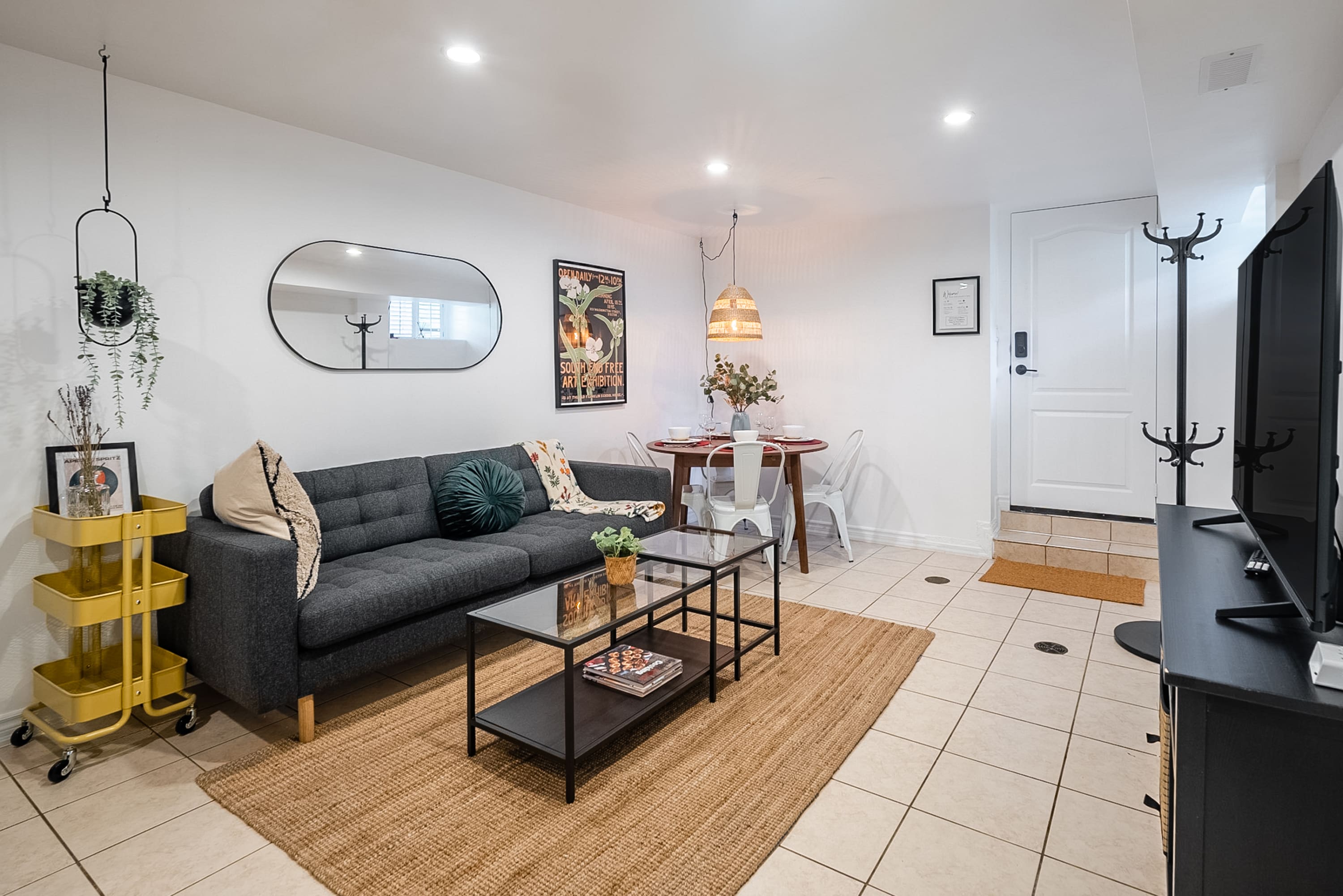 The living room features a dark gray sofa, a glass-top coffee table on a jute rug, and a round dining table with white chairs. A black TV stand with a large flat-screen TV is positioned near the entrance, which has a white door and a coat rack.