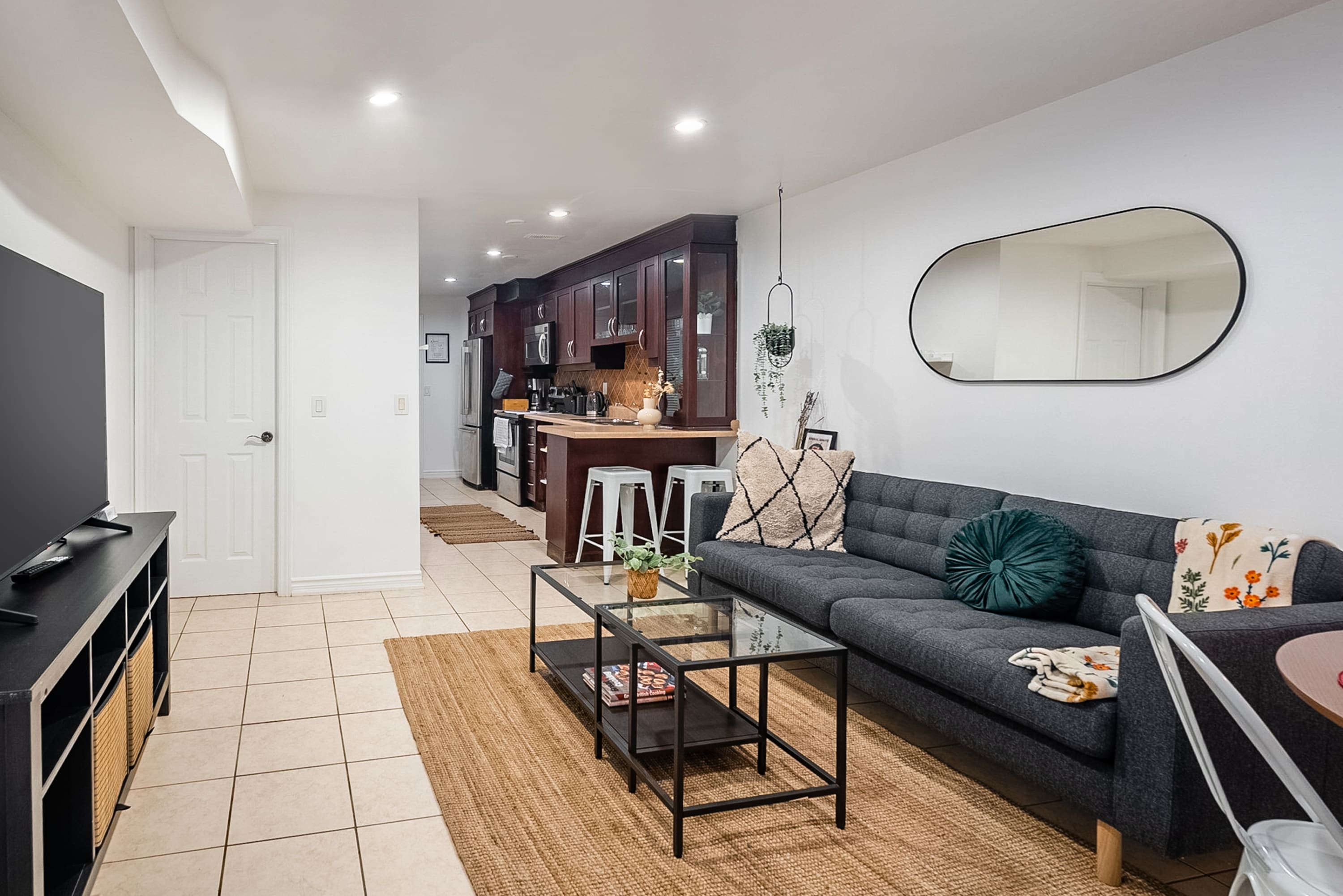 A view from the living room shows a dark gray sofa and glass coffee table, with a black TV stand and flat-screen TV on the left. The tiled floor extends towards the kitchen with dark wood cabinets and a breakfast bar, and a white door is visible in the background.