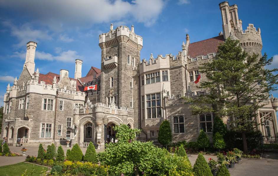A majestic, historic stone castle with multiple towers and intricate details, set against a bright blue sky with fluffy clouds. The castle is surrounded by meticulously manicured green lawns and various shrubs.