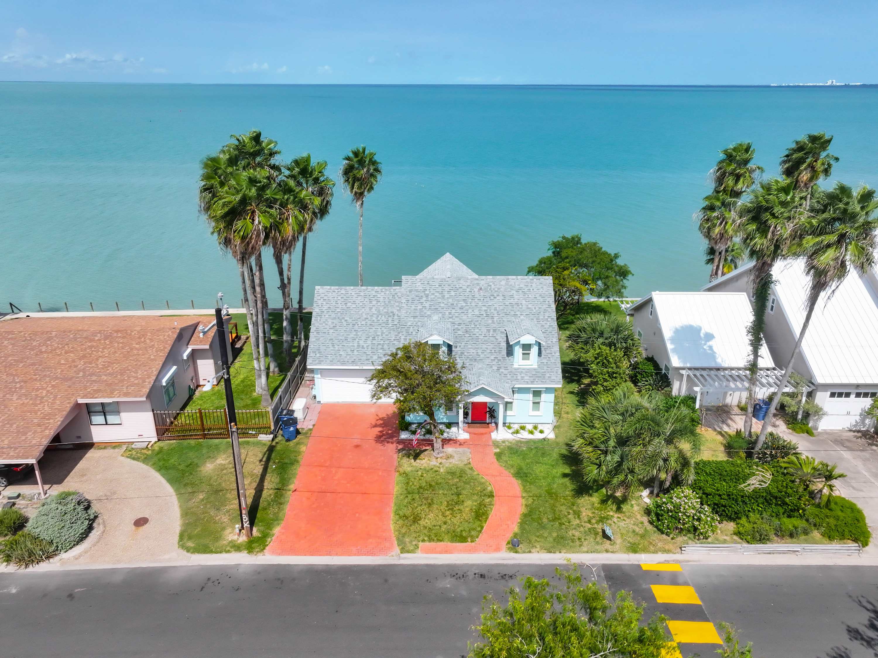 A direct aerial shot highlights the light blue house with its red driveway and pathway, positioned directly on the edge of the serene, turquoise bay. The property is surrounded by green lawn and palm trees, emphasizing its prime waterfront location.