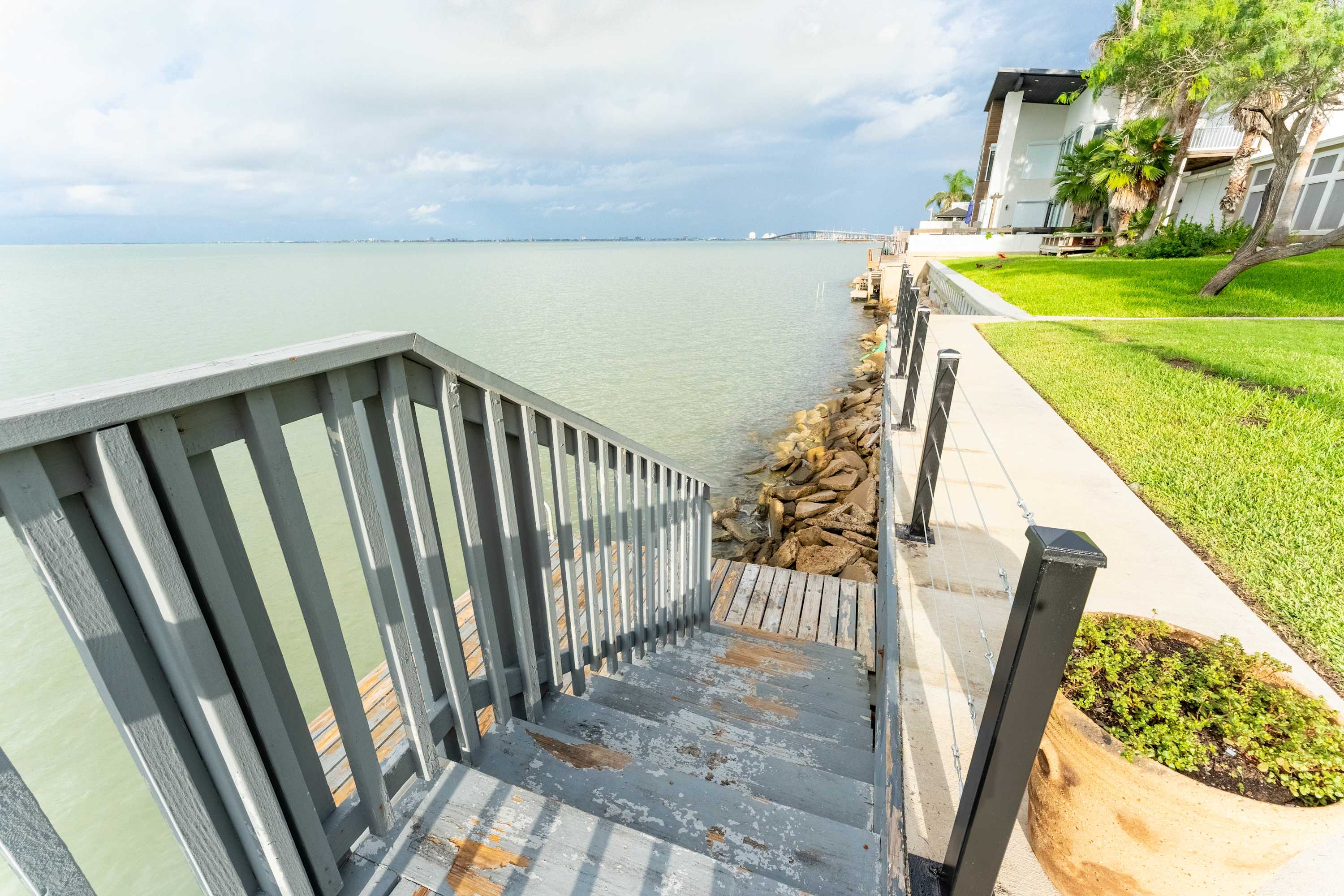 Weathered wooden stairs with grey railings descend towards the calm bay water, with a rocky shoreline and a modern building visible in the distance. Lush green grass and potted plants frame the top of the stairs.