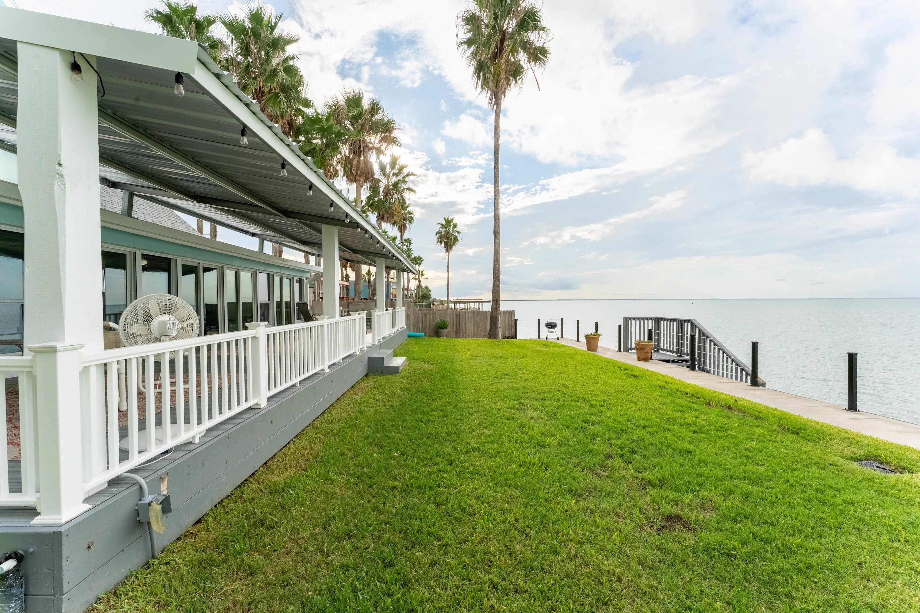 The back exterior of the house features a long covered patio with white railings, overlooking a gently sloping green lawn that leads directly to the bay. Tall palm trees line the property, enhancing the tropical waterfront ambiance.