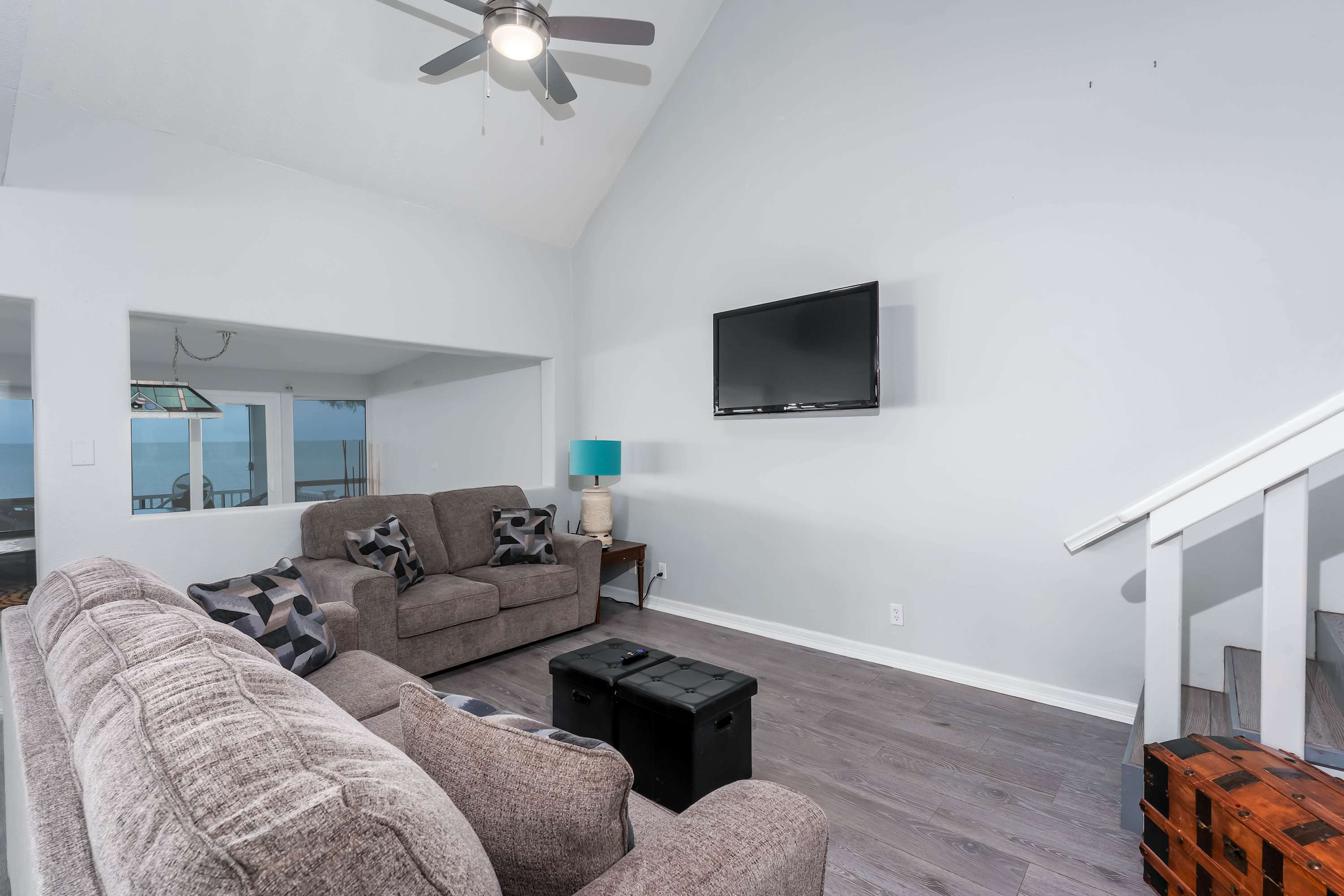 A bright living room with light gray walls and light wood-look flooring, featuring two taupe sofas and a wall-mounted TV. A large opening in the wall provides a view of the water, and a staircase is visible on the right.