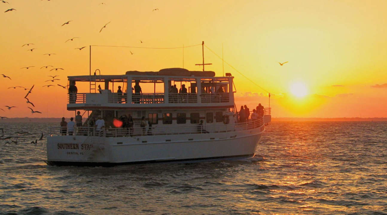 A large passenger boat, 'Southern Star,' cruises on the water during a stunning sunset, casting a warm orange glow across the sky. Numerous birds fly overhead, adding to the picturesque and serene evening scene.