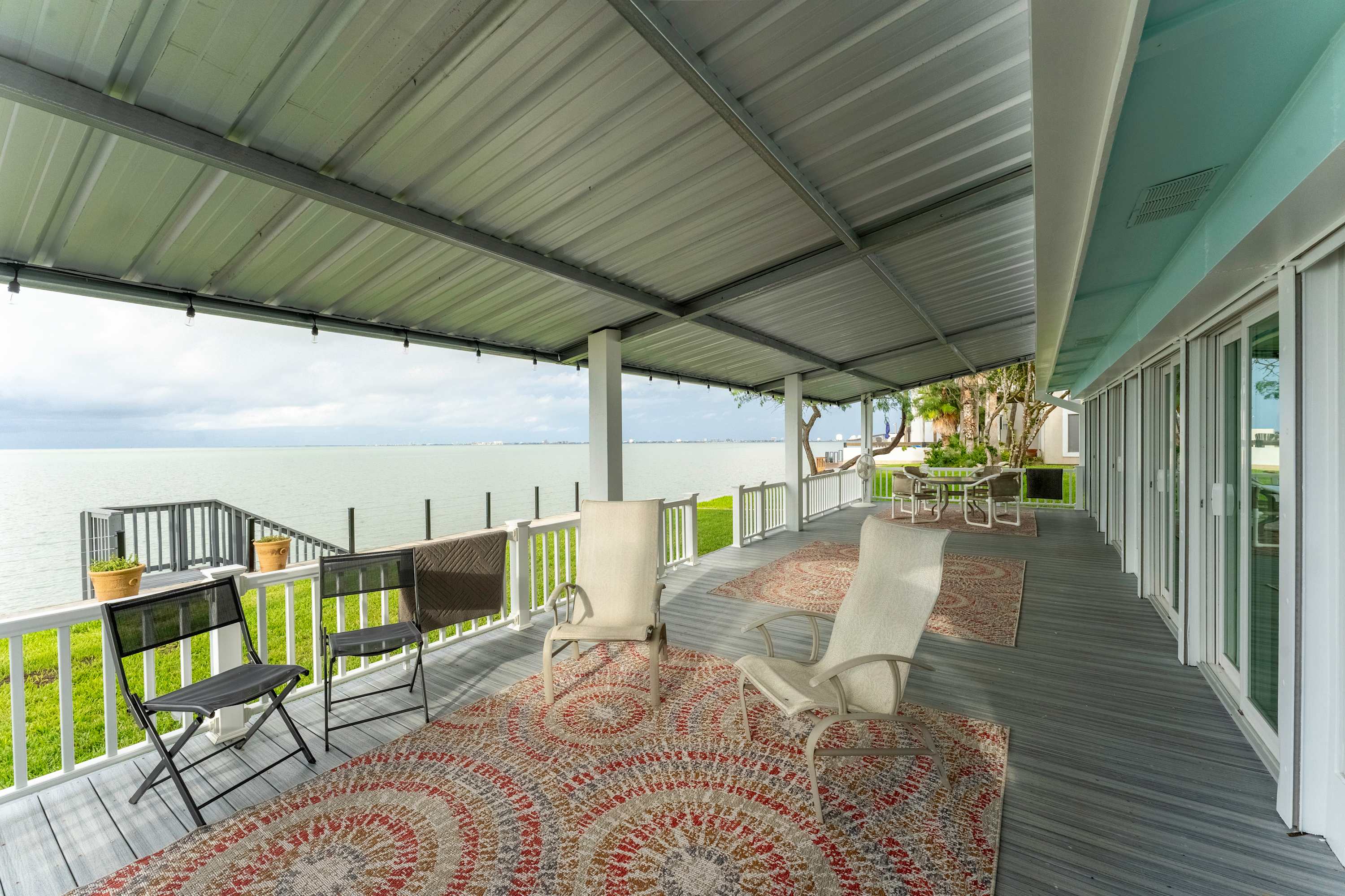A long, covered outdoor deck with gray flooring and white railings, overlooking the bay. Multiple outdoor rugs and various chairs are arranged along the deck, providing several seating areas with water views.