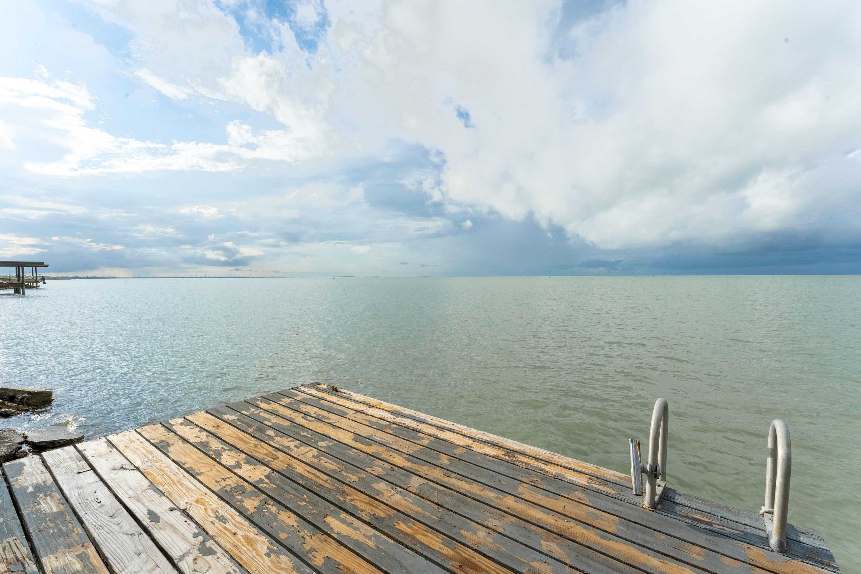 A rustic wooden dock extends into the expansive, calm bay under a partly cloudy sky, featuring metal ladders for easy water access. The weathered planks show signs of age, adding character to the waterfront setting.