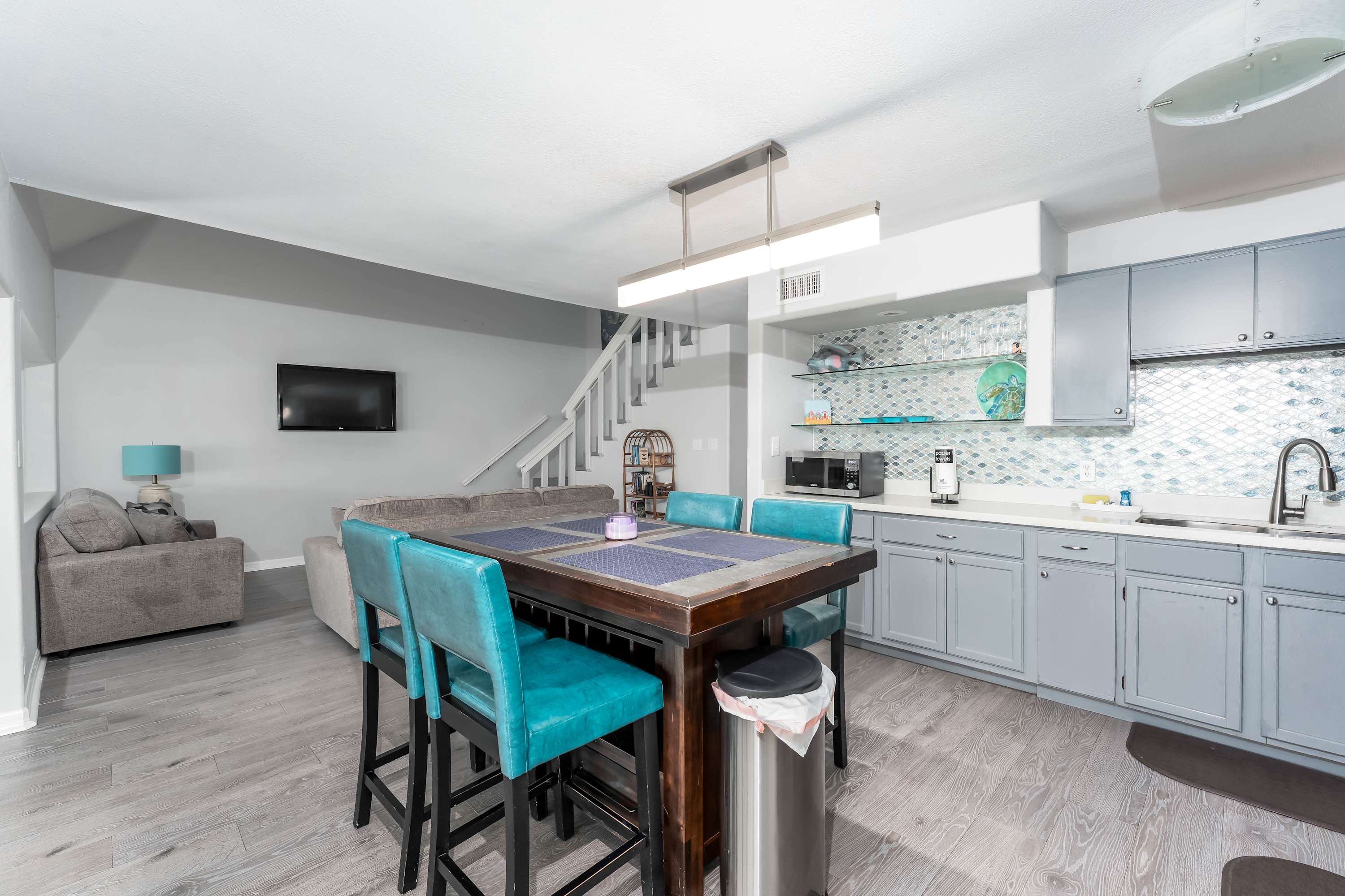 A dining area with a large wooden table and teal chairs, adjacent to a kitchen area with gray cabinets, white countertops, and a tiled backsplash. The space has light wood-look flooring and is open to the living room.