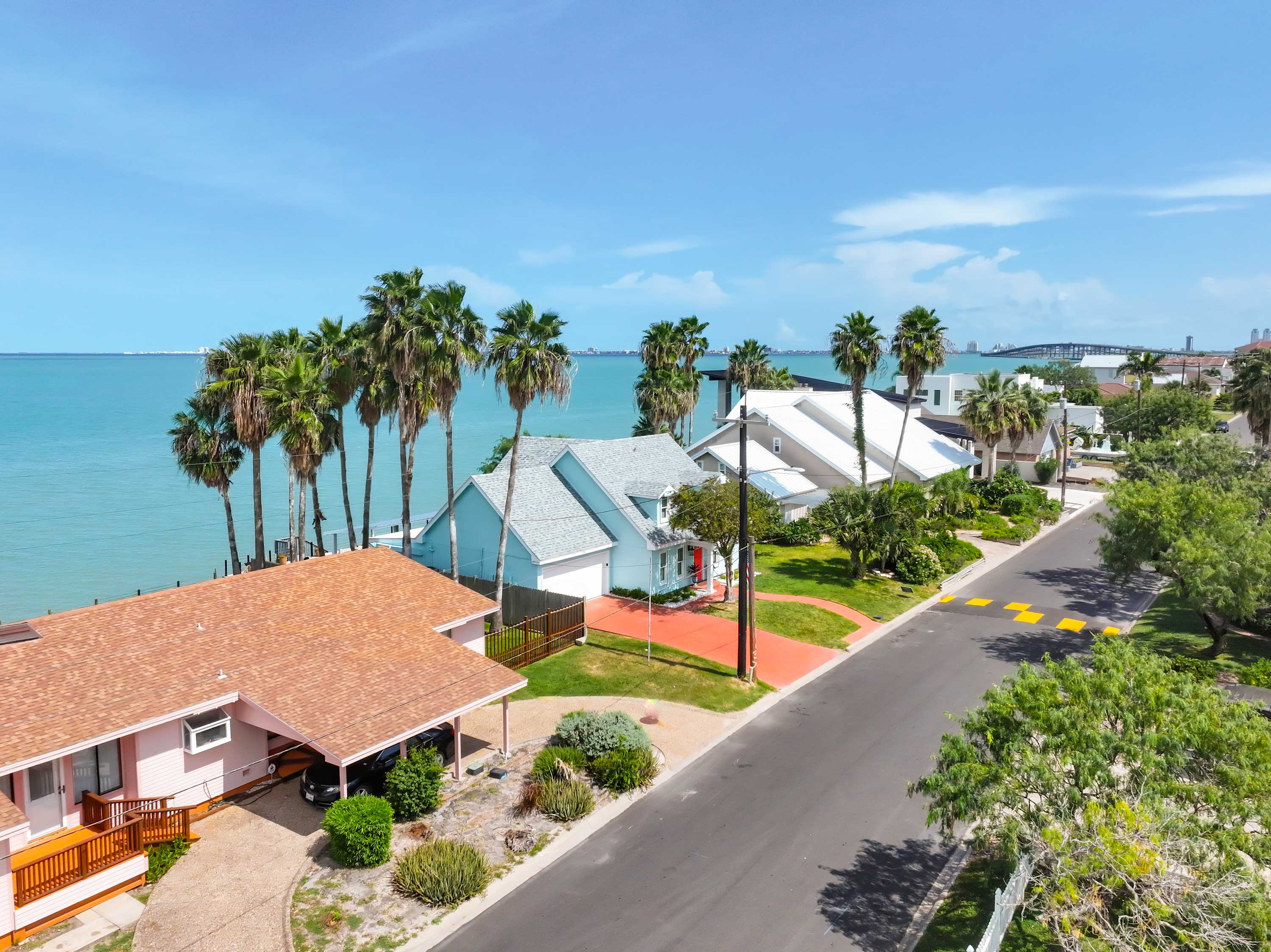 An aerial view showcases a residential street lined with houses, including the light blue subject property, all situated along the vibrant turquoise bay. Palm trees dot the landscape, and a distant bridge is visible across the water.